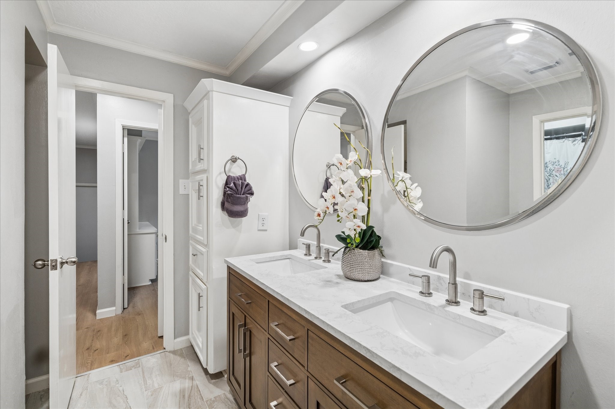 This bathroom features a modern double vanity with a sleek marble countertop, large round mirrors, and contemporary fixtures. It has ample storage with a tall cabinet and stylish wood cabinetry, set against a neutral color palette.