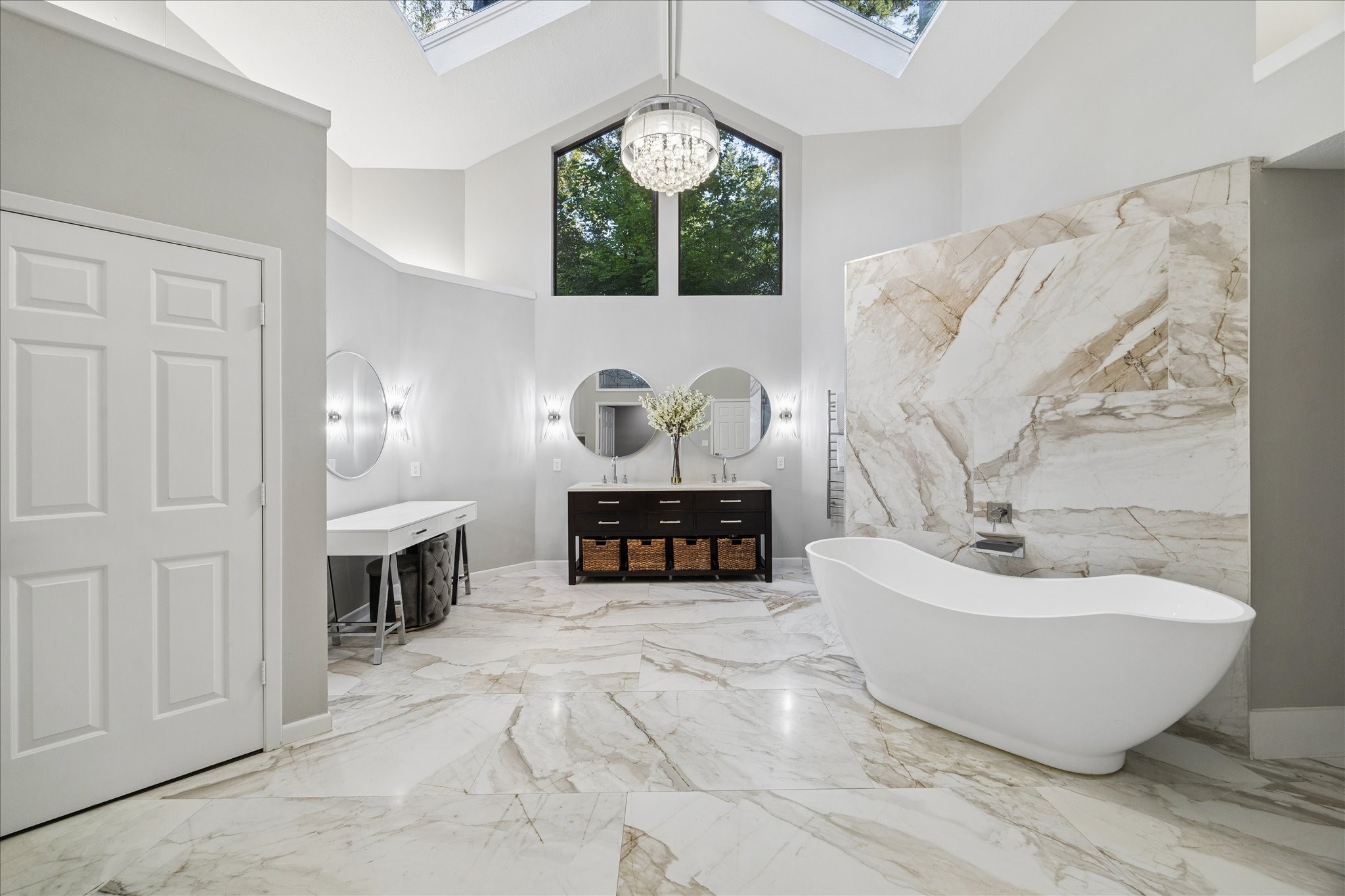This luxurious primary bathroom features elegant marble flooring and walls, a freestanding bathtub, and a large window that fills the space with natural light. The room is complemented by a modern vanity with double sinks, two round mirrors, and a chandelier for added sophistication.