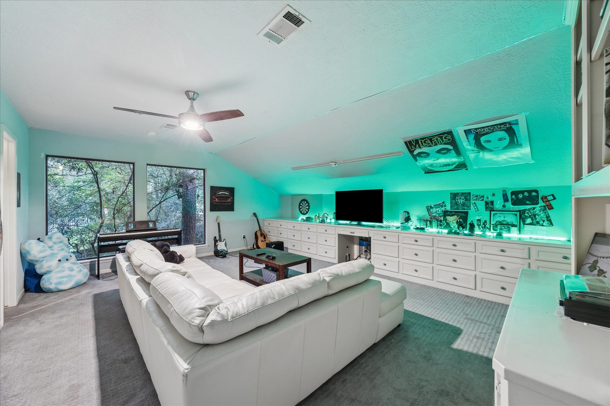 This 2nd-floor spacious entertainment room features a cozy white sectional, ambient lighting, and a wall-mounted TV. It includes ample storage with built-in cabinets and large windows providing natural light and views of the backyard.