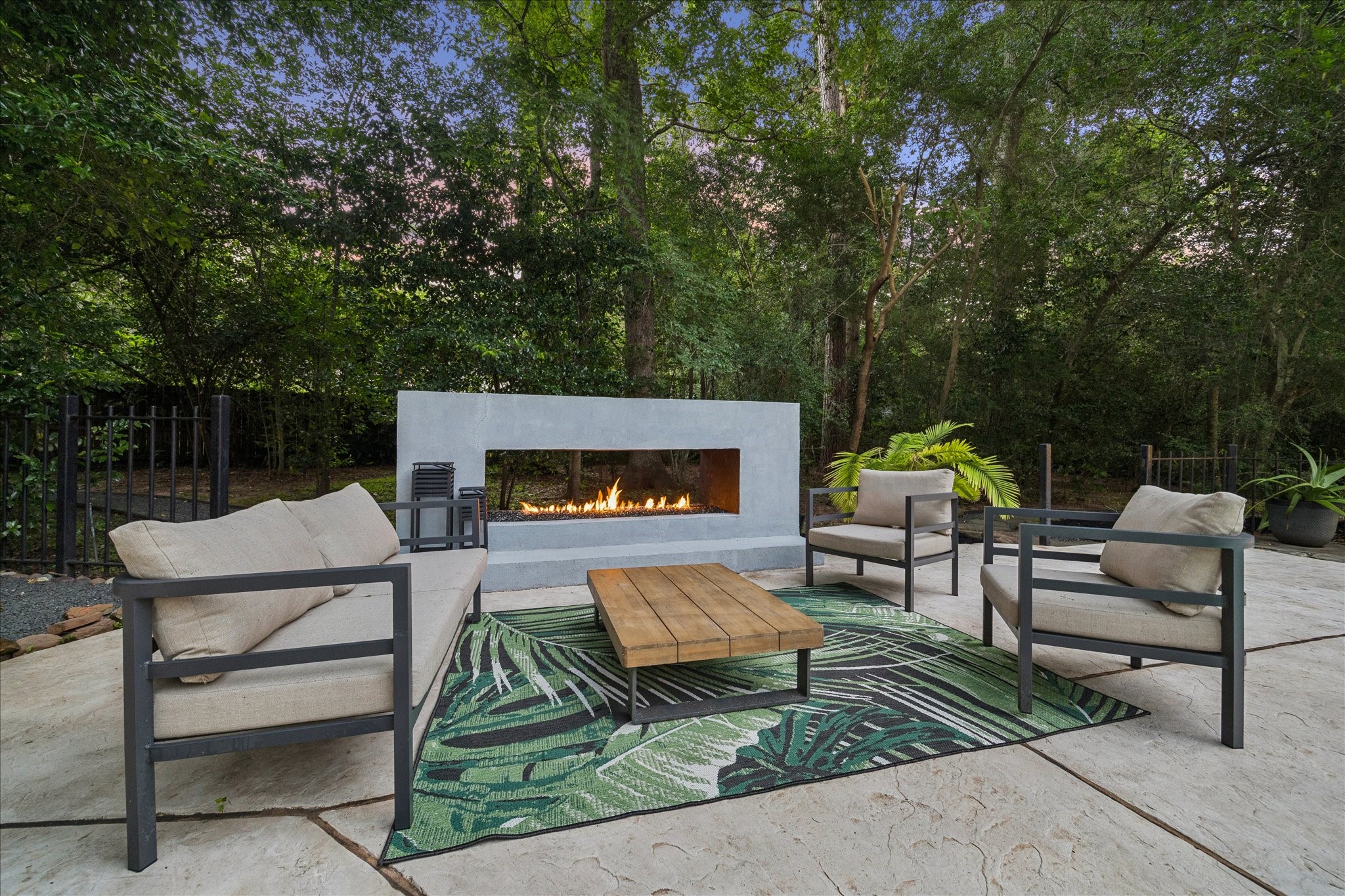 This inviting outdoor space features a modern fire pit surrounded by comfortable seating, set on a stylish patio with a lush, wooded backdrop. Perfect for relaxing or entertaining.