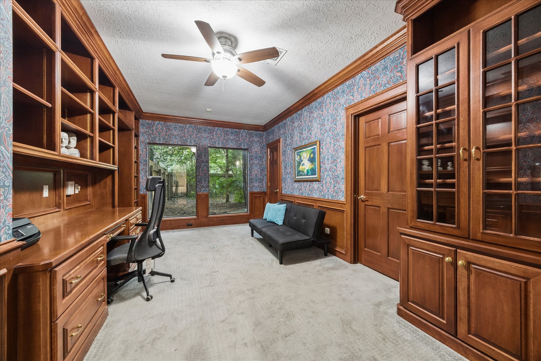 This is the 2nd home office next to the primary suite, with rich wood built-in shelves and cabinets. It features a large desk area, a comfortable chair, and a sofa. Two large windows provide natural light and a view of greenery, while a ceiling fan adds comfort. The room has a classic, elegant feel with its wood details and patterned wallpaper.