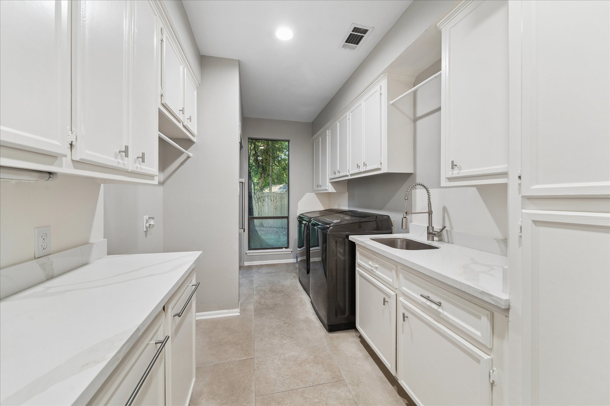 The laundry room features sleek white cabinetry, ample storage, and counter space. It features a utility sink and a window providing natural light. The neutral tile flooring complements the clean, bright aesthetic.