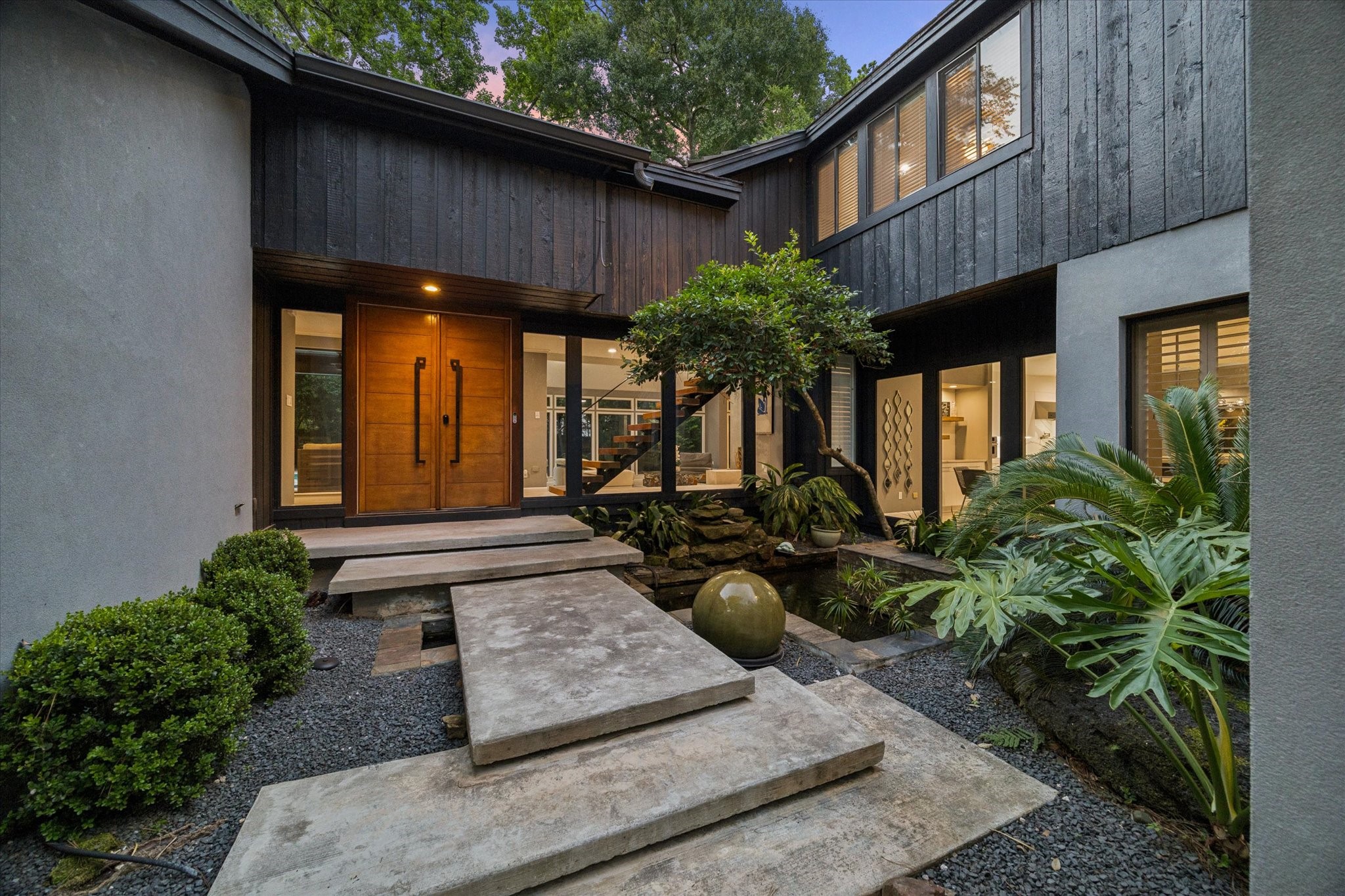Stunning entrance featuring a minimalist stone pathway over a serene water feature and koi pond framed by lush greenery and sleek, dark wood paneling. Large windows provide a glimpse into the inviting interior, creating a harmonious blend of nature and architecture. Stunning entrance featuring a minimalist stone pathway over a serene water feature and koi pond framed by lush greenery and sleek, dark wood paneling. Large windows provide a glimpse into the inviting interior, creating a harmonious blend of nature and architecture.