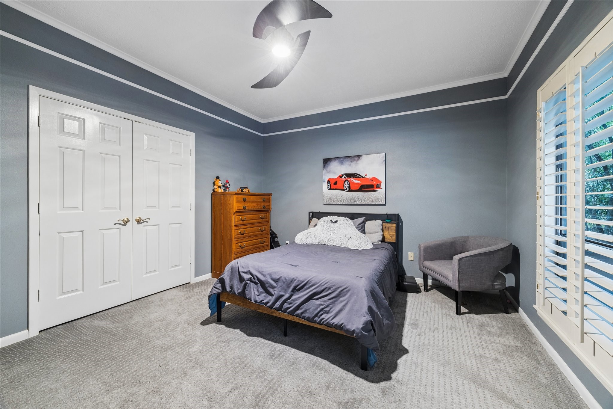 The first-floor guest bedroom features a cozy atmosphere with gray walls, a carpeted floor, and a ceiling fan. Natural light enters through large shuttered windows, and there's a closet with double doors for ample storage.