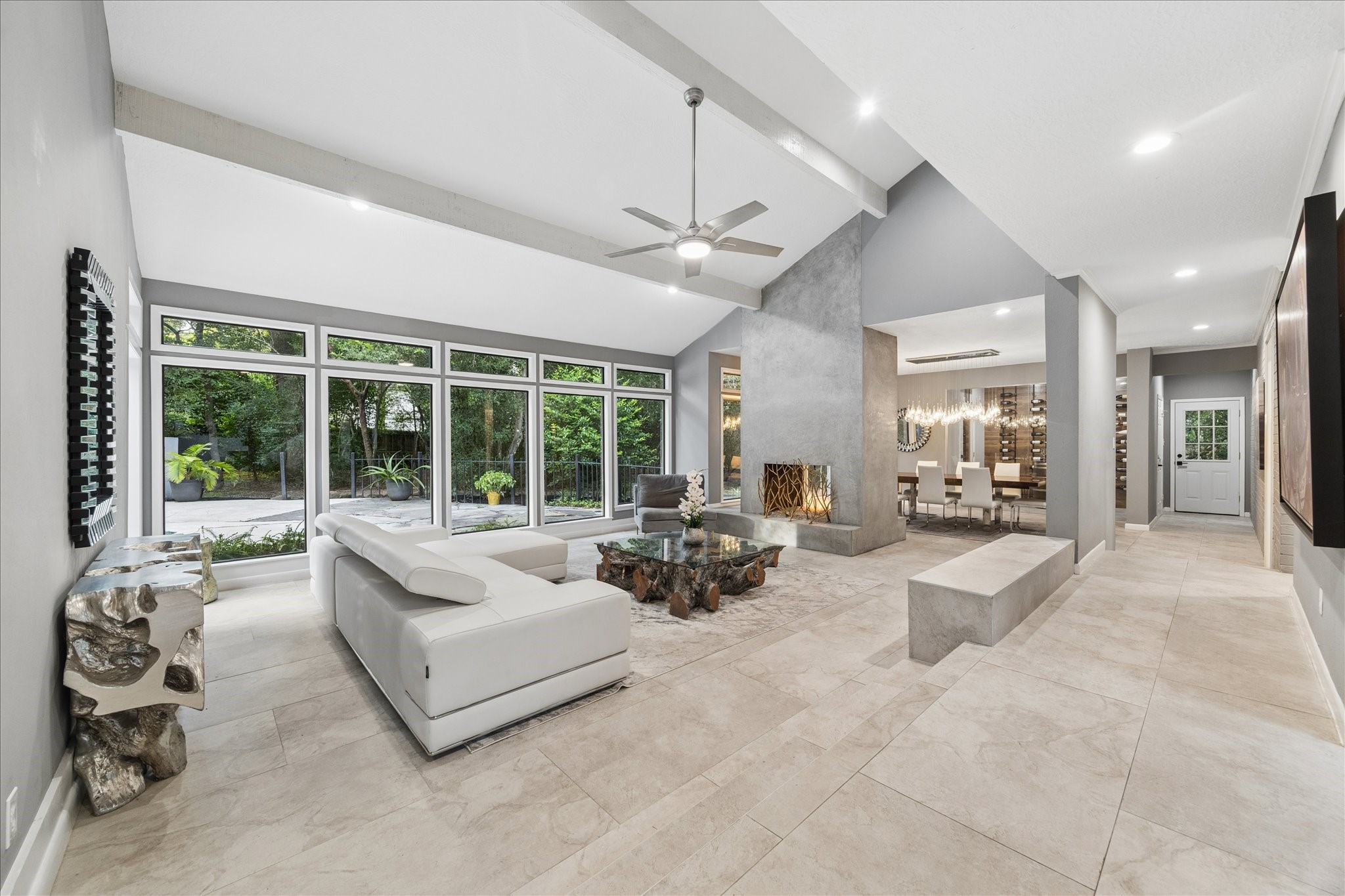 This modern living room features a sleek design with large floor-to-ceiling windows offering ample natural light and a view of lush greenery. It includes a contemporary fireplace, elegant dining area, and neutral-toned flooring, creating a spacious and inviting atmosphere.