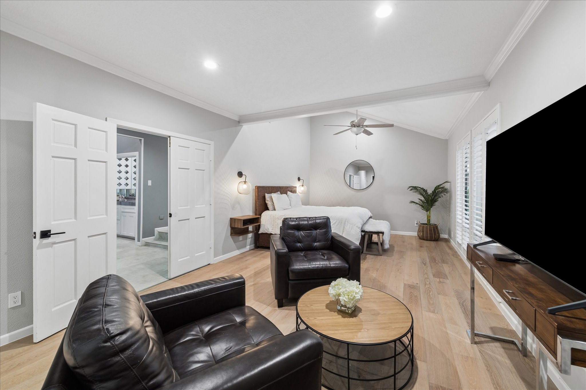 This spacious primary suite features a cozy seating area with leather chairs, a large flat-screen TV, and a stylish round coffee table. The bed area is well-lit with wall-mounted lamps, and the room has wood flooring, high ceilings with a fan, and ample natural light from the windows.