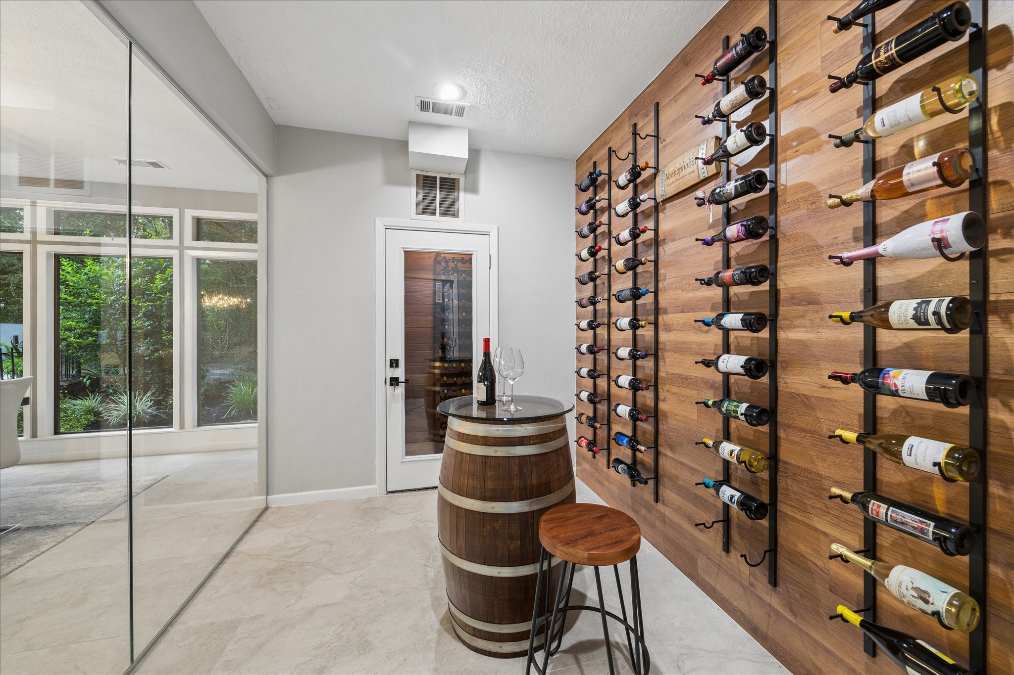 Enjoy your very own wine cellar and climate-controlled wine room. With a stylish wooden wall-mounted wine rack, a glass door, and a cozy tasting area featuring a barrel table and stool. Large windows provide a view of lush greenery, adding a serene backdrop.