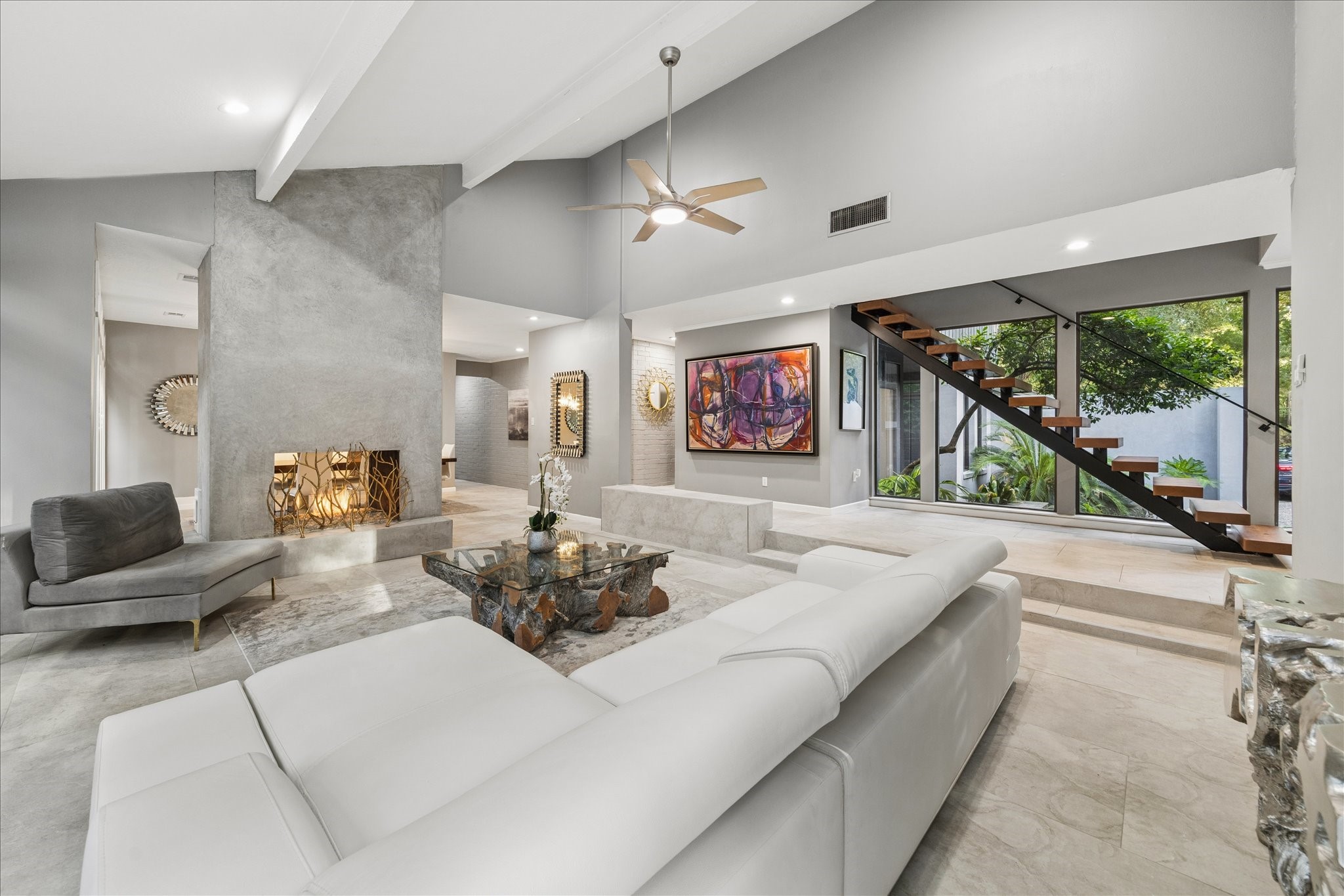 This spacious, modern living room features high ceilings, a sleek, open staircase, and large windows offering abundant natural light. The contemporary design includes a textured fireplace, elegant gray and white tones, and stylish furnishings, creating a welcoming and sophisticated atmosphere.