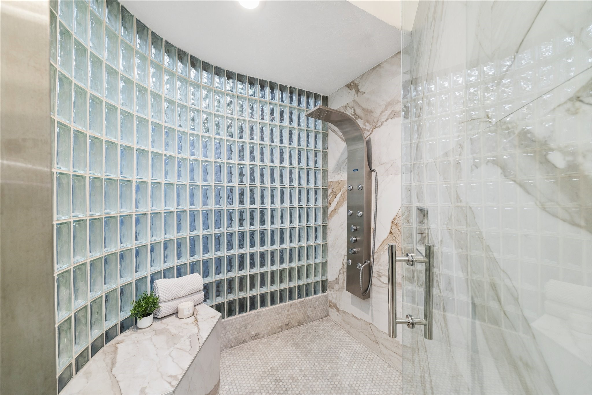 The primary bathroom features a spacious glass block shower with elegant marble walls and floors. It includes a sleek shower panel with multiple settings, complemented by a built-in marble bench for added comfort and luxury.
