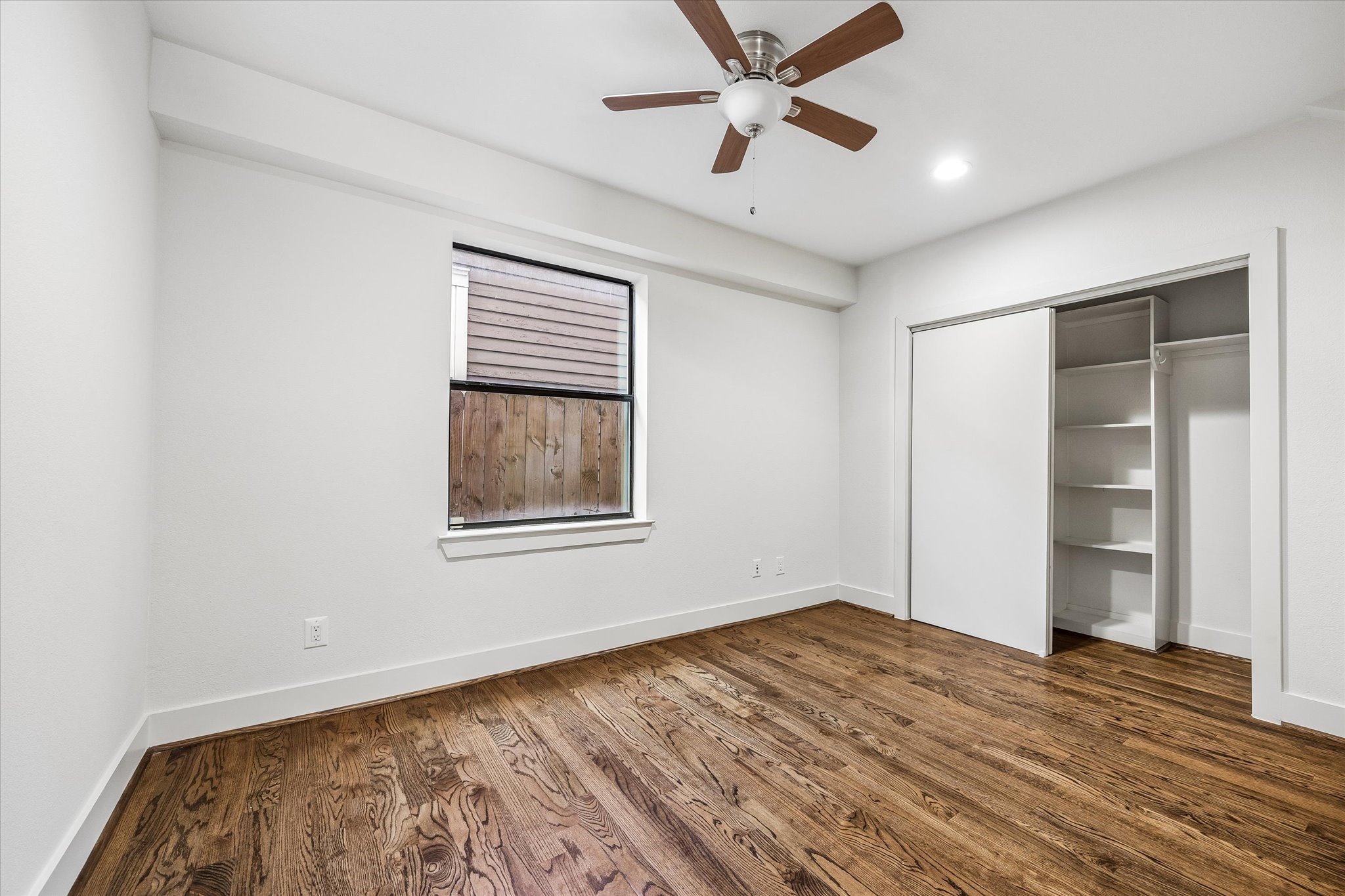 First floor bedroom with wood flooring, a ceiling fan, and a window for natural light. A wide closet includes built-in shelving and hanging space behind sliding doors.