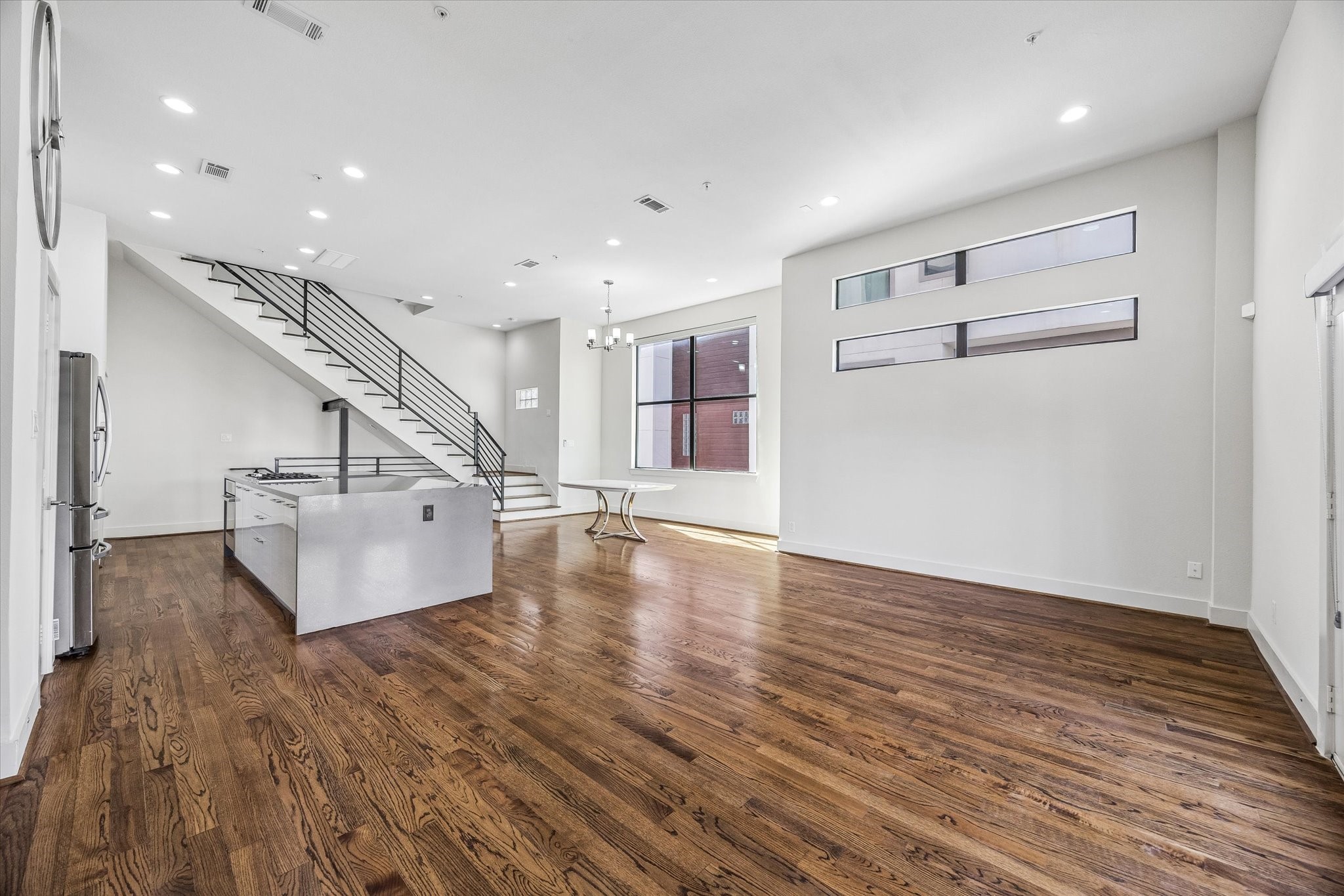 Open living, dining, and kitchen areas offer a connected layout with abundant natural light, continuous wood flooring, and a staircase with horizontal metal railings. Open living, dining, and kitchen areas offer a connected layout with abundant natural light, continuous wood flooring, and a staircase with horizontal metal railings.