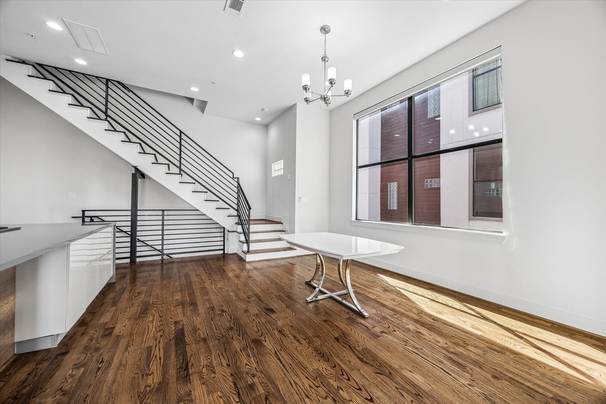 Bright dining area beside an oversized window, with wood flooring and an open connection to the kitchen. The adjacent staircase features wood treads and horizontal metal railings.