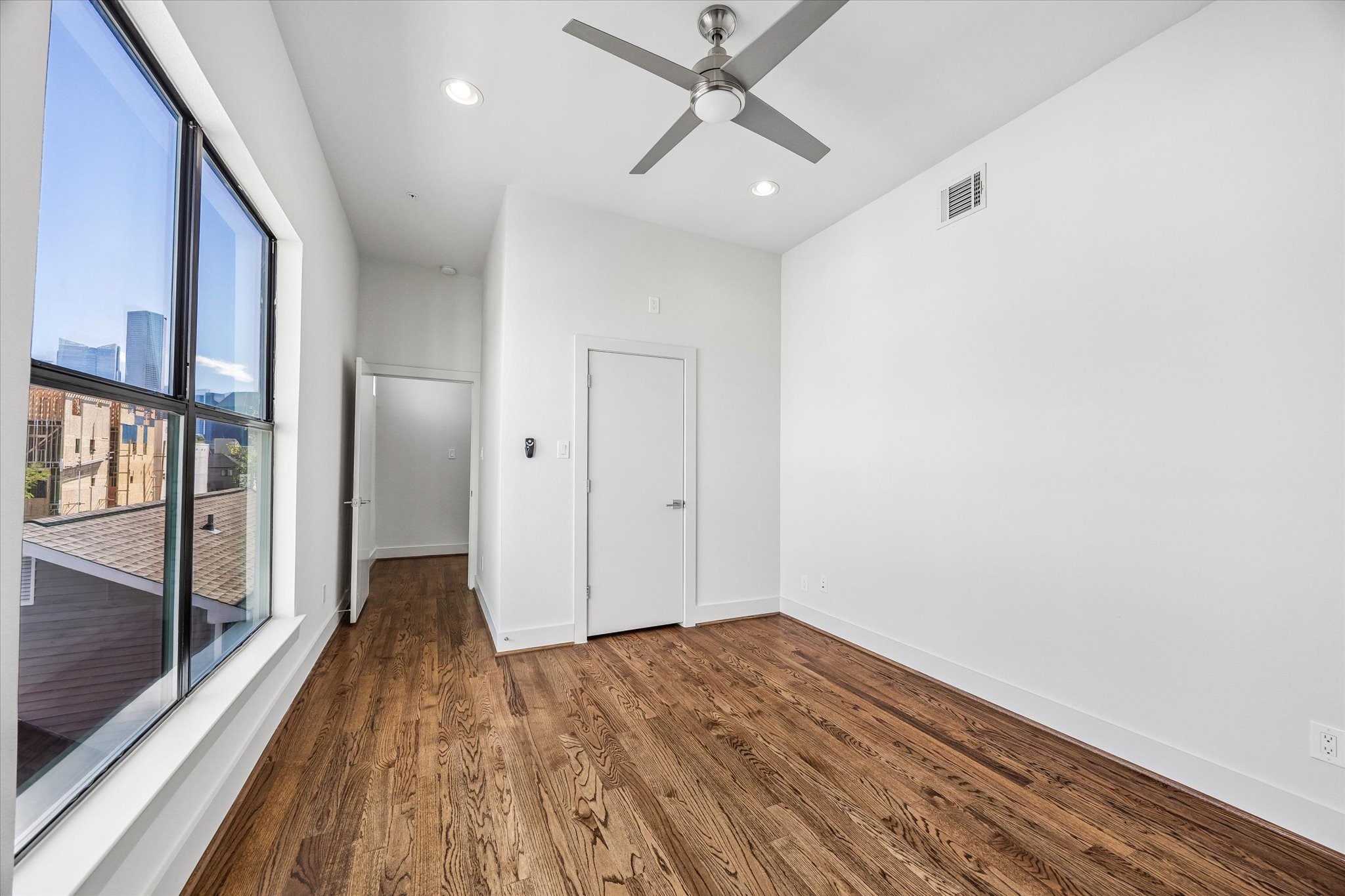 Alternate bedroom view showcasing the large window with a glimpse of the city skyline, along with wood flooring, a ceiling fan, and closet access.