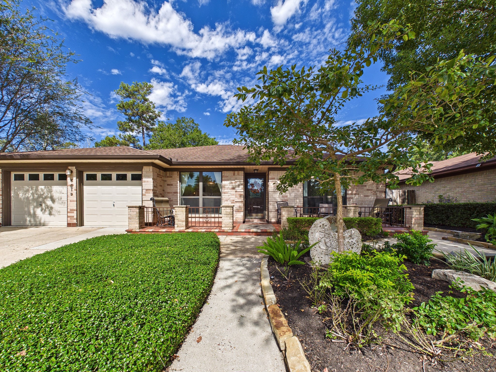 Charming brick home with a welcoming front porch, lush landscaping, and a spacious driveway leading to a double garage.
