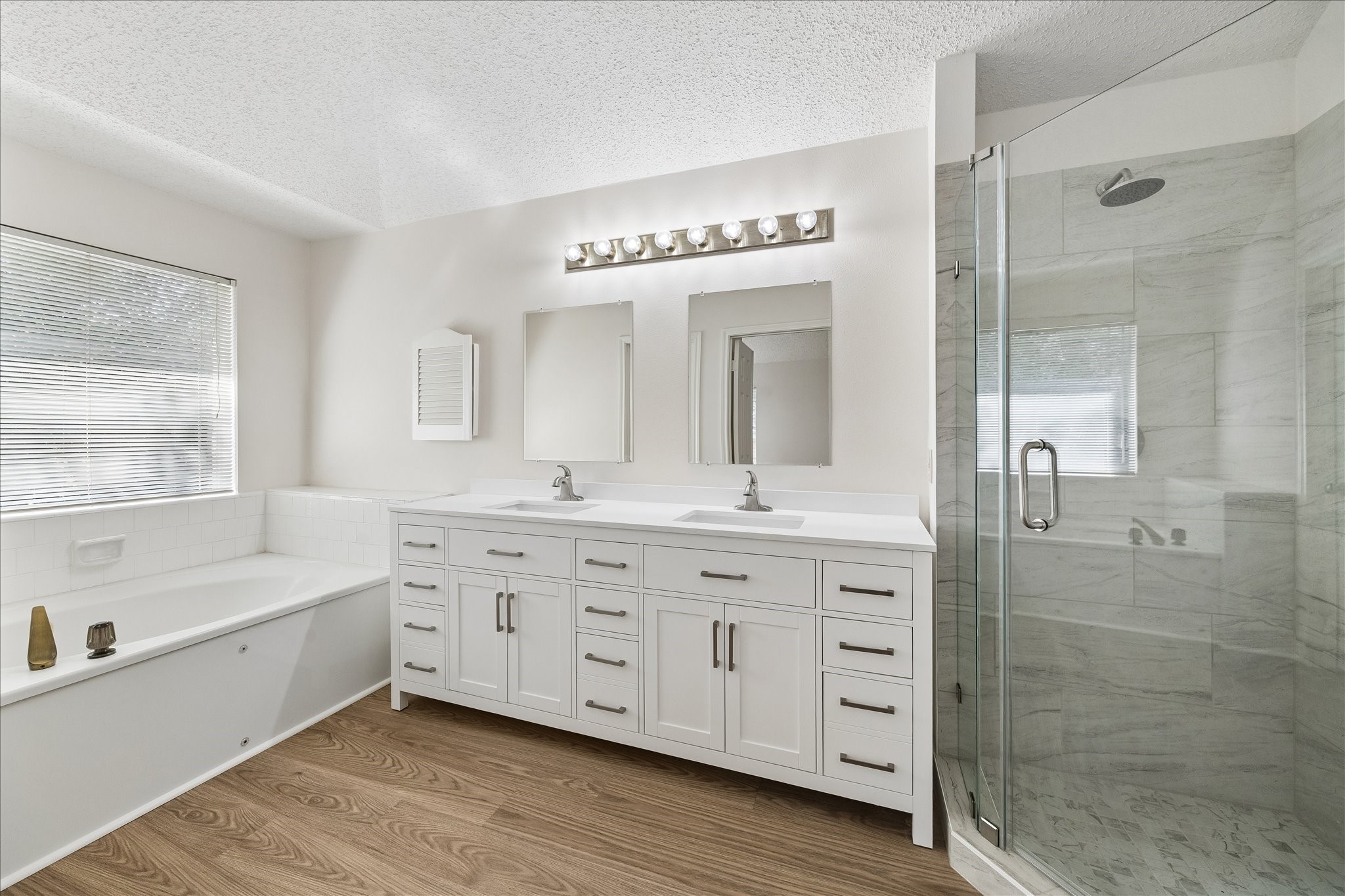 Tastefully remodeled primary bathroom with new flooring, cabinets, double sinks, mirrors and glass enclosed shower.