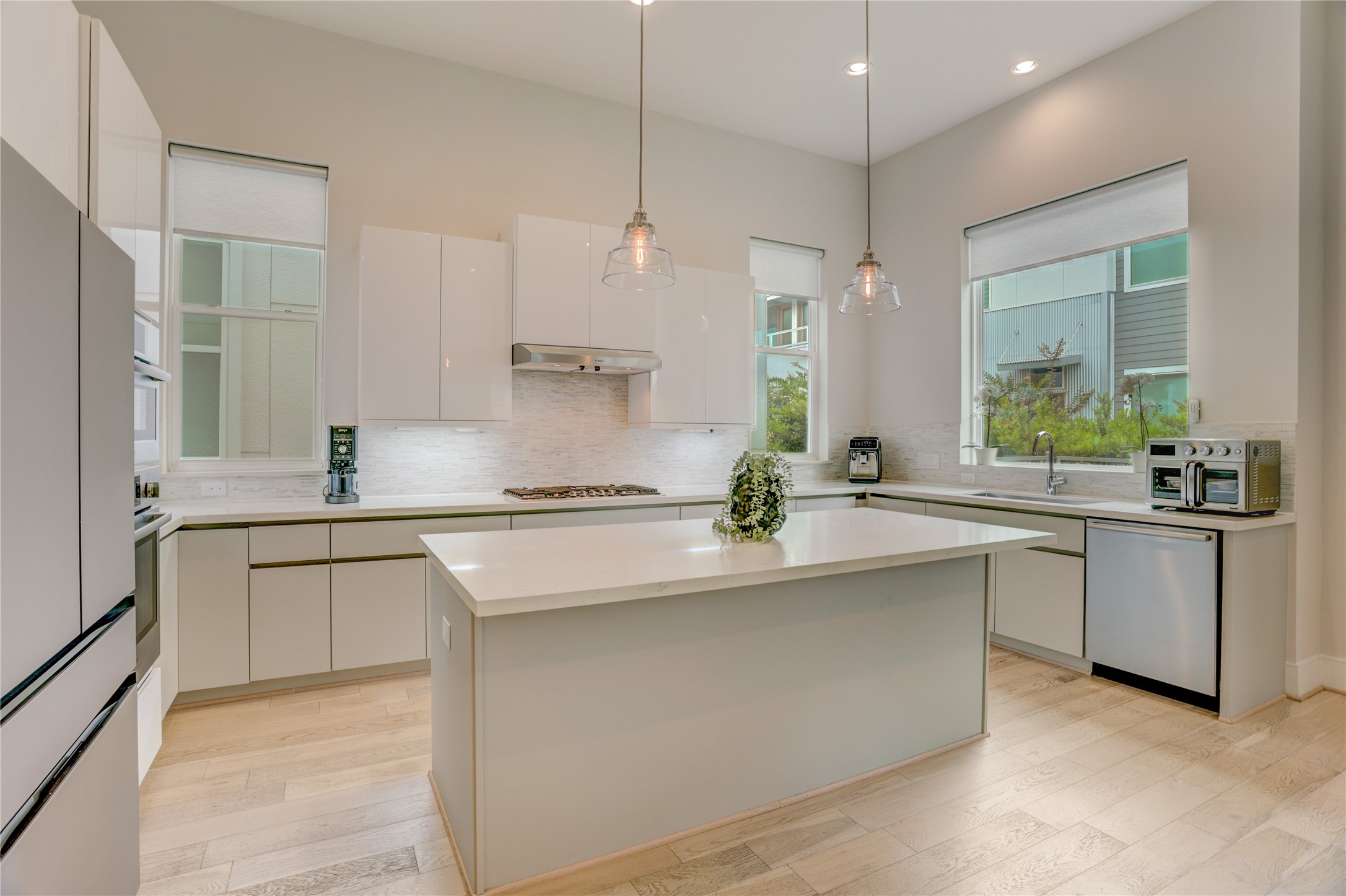 Modern kitchen with sleek white cabinetry, a spacious island, and stainless steel appliances, illuminated by stylish pendant lighting and natural light from large windows.
