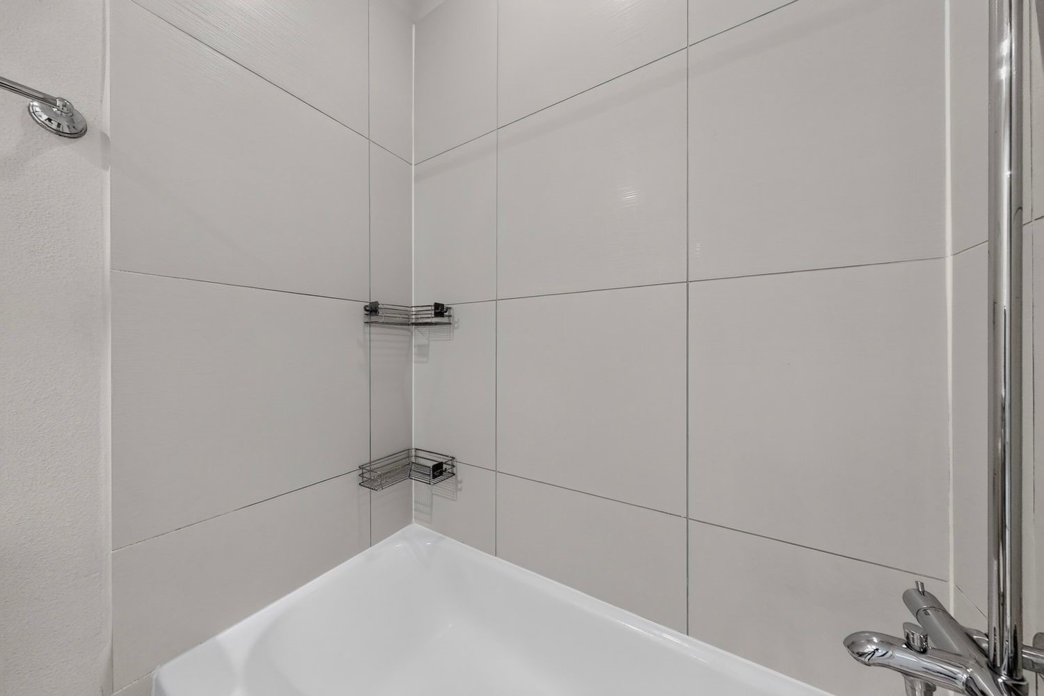 The Sleek and modern shared guest bathroom with large tiles, a chrome showerhead, and convenient corner shelving for storage.