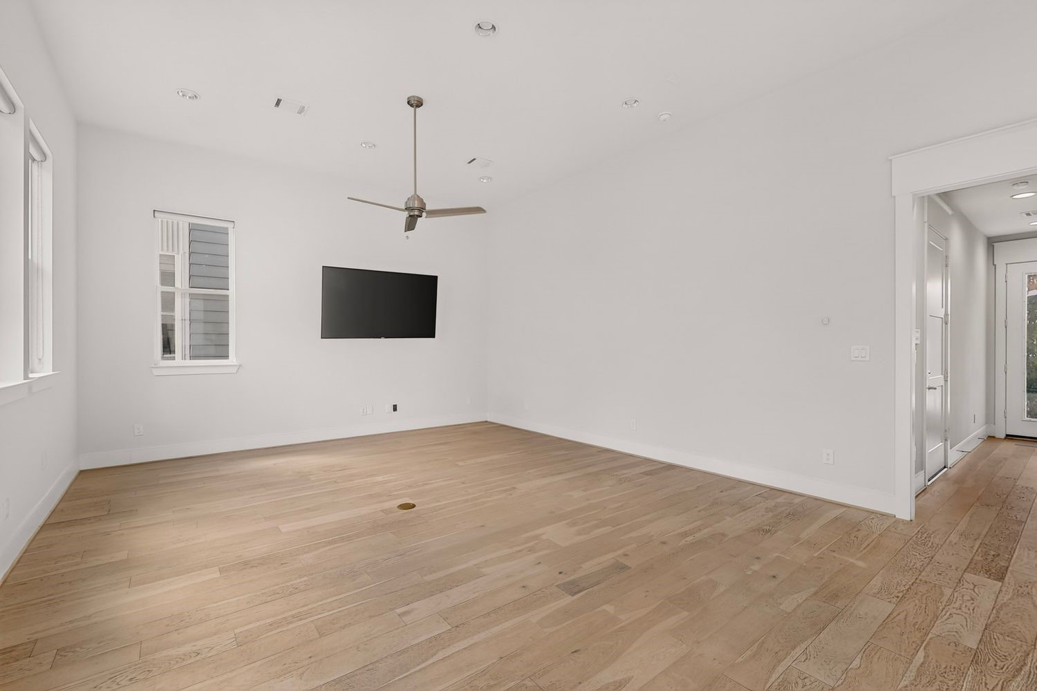 The vacant look at the Spacious living area with elegant hardwood floors, a modern ceiling fan, and a mounted TV, offering ample natural light through multiple windows.