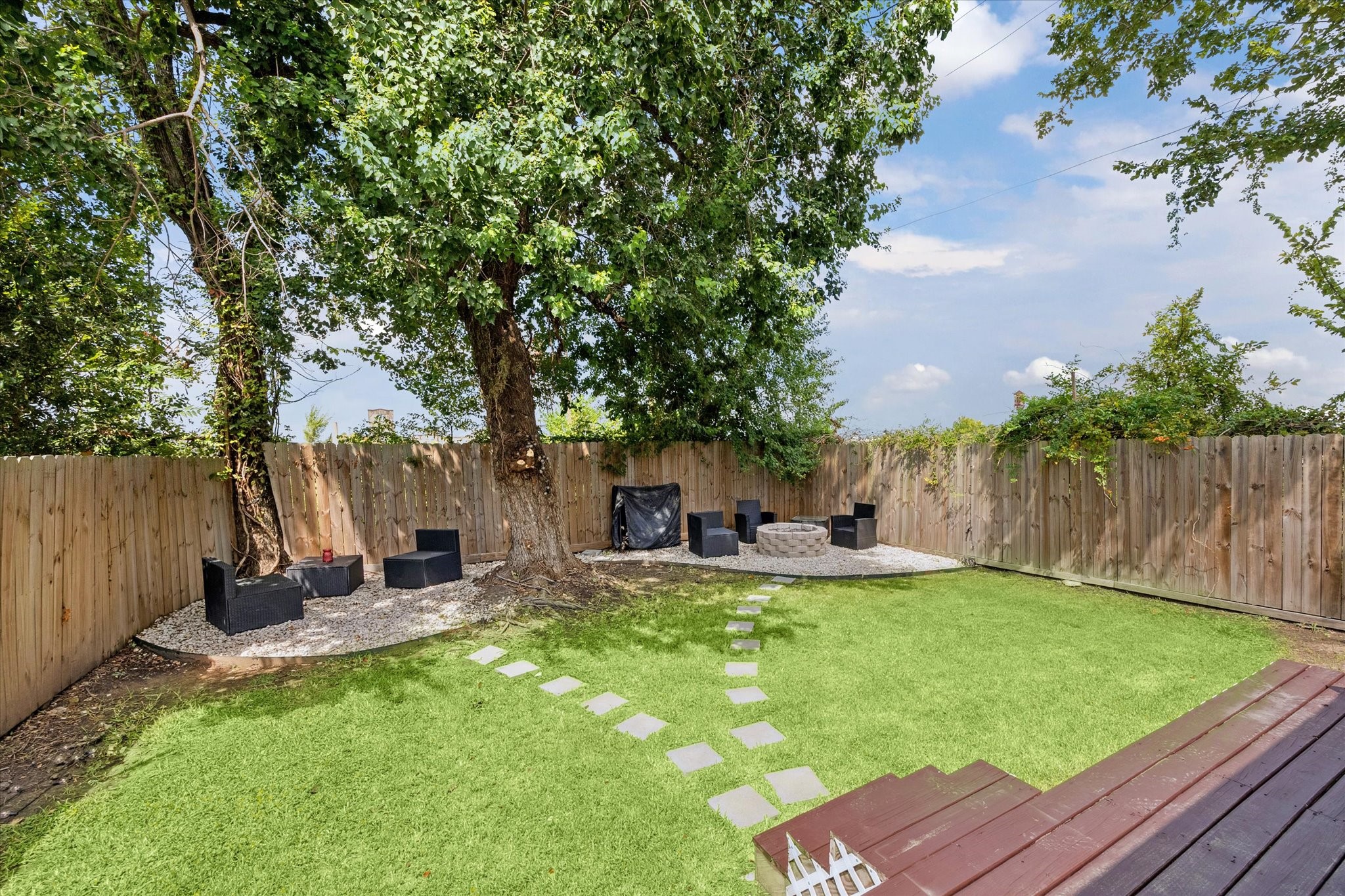 Mature shade trees frame a private backyard retreat with multiple seating areas, including a firepit corner perfect for evening gatherings. The fully fenced yard combines green space with low-maintenance gravel accents for easy enjoyment.