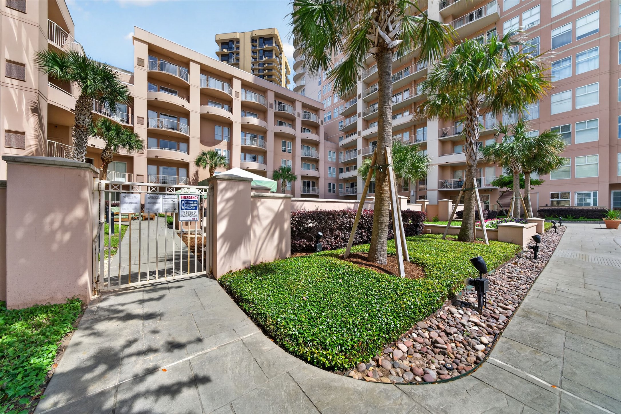 Modern condominium complex with lush landscaping, palm trees, and a secure gated entrance, offering a welcoming and stylish living environment.