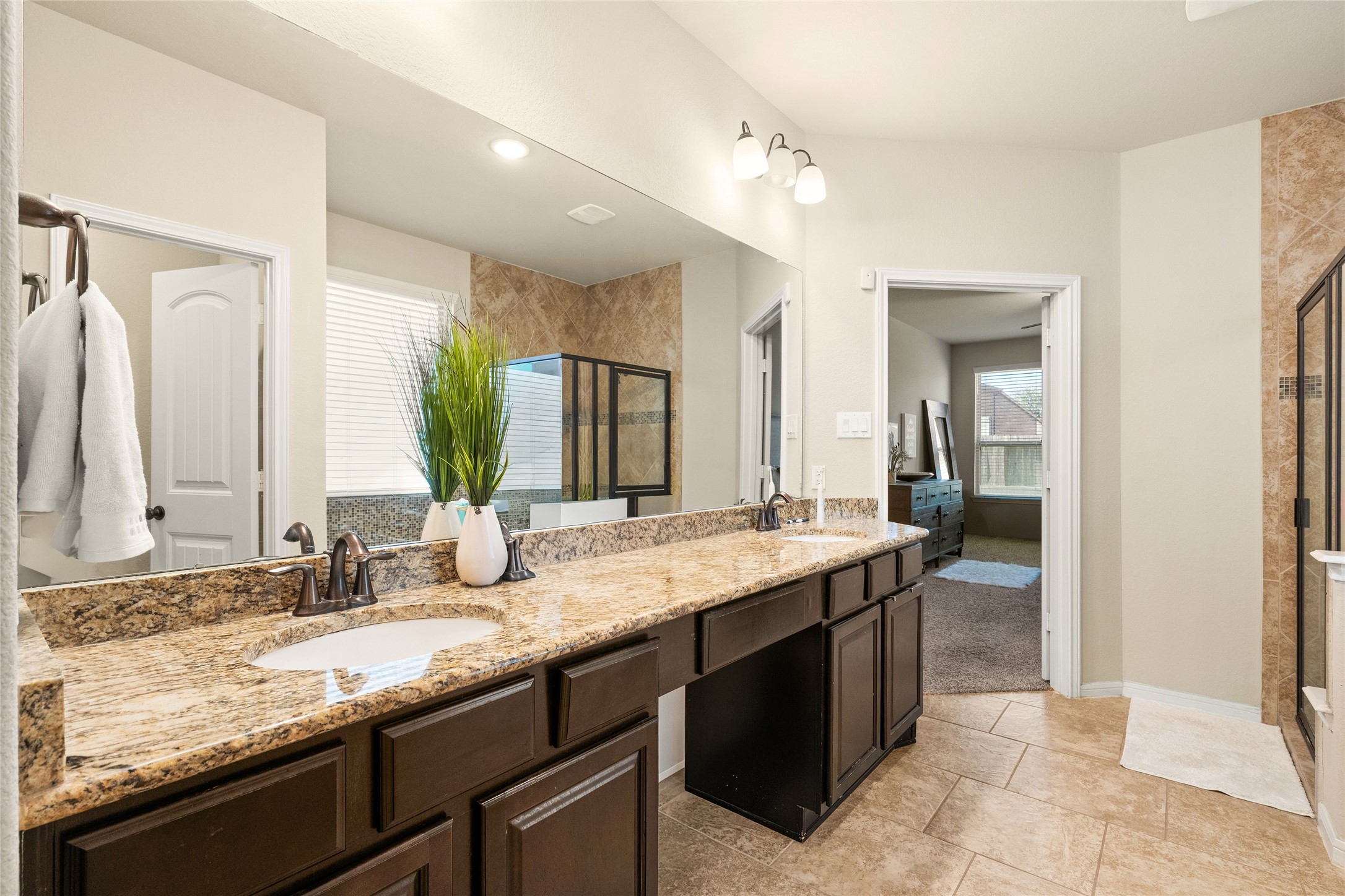 Spacious bathroom featuring a double vanity with granite countertops, elegant lighting, and a walk-in shower with glass doors, seamlessly connected to the master bedroom.