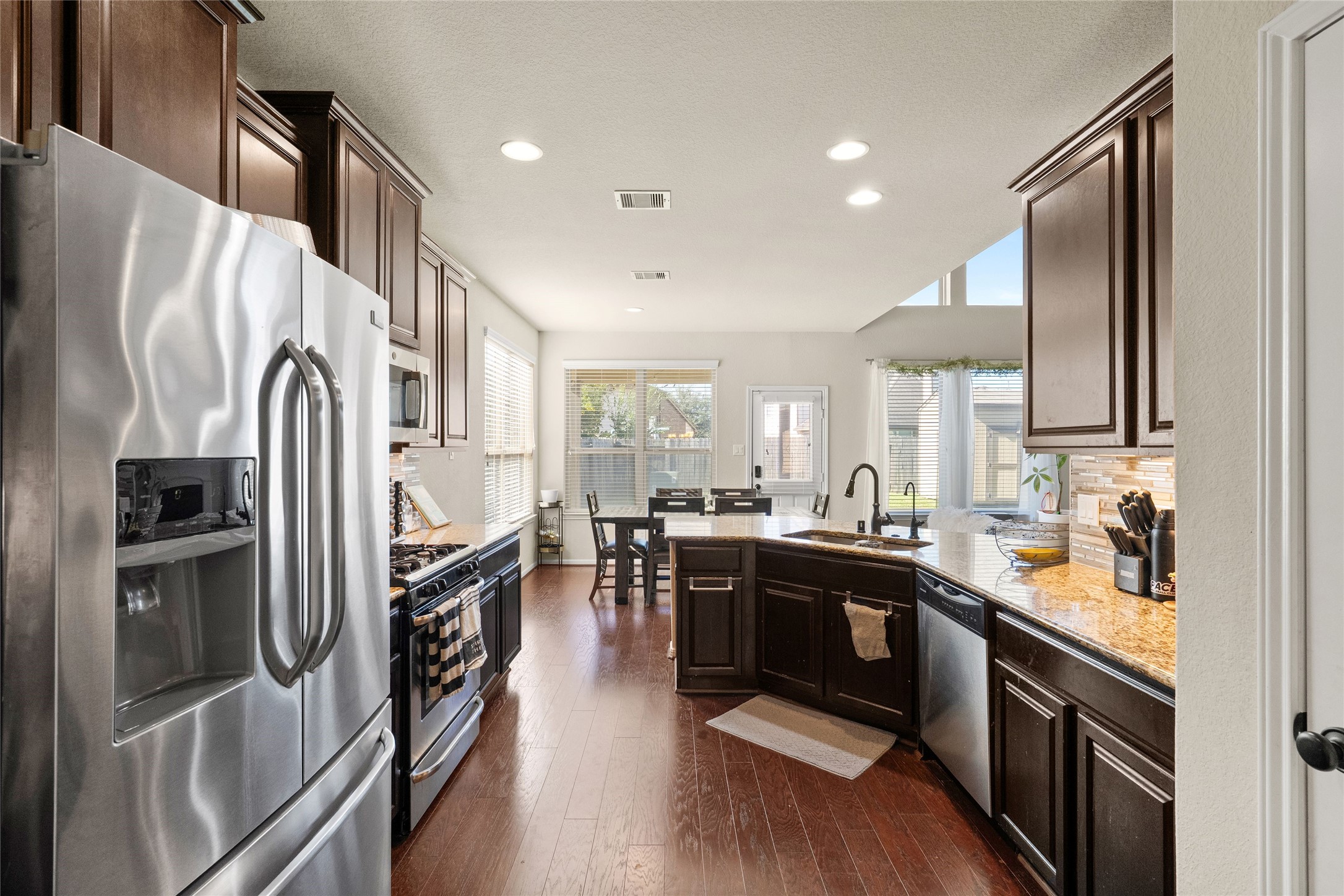 Spacious kitchen with rich dark cabinetry, stainless steel appliances, and granite countertops, leading to a bright dining area with large windows and natural light.