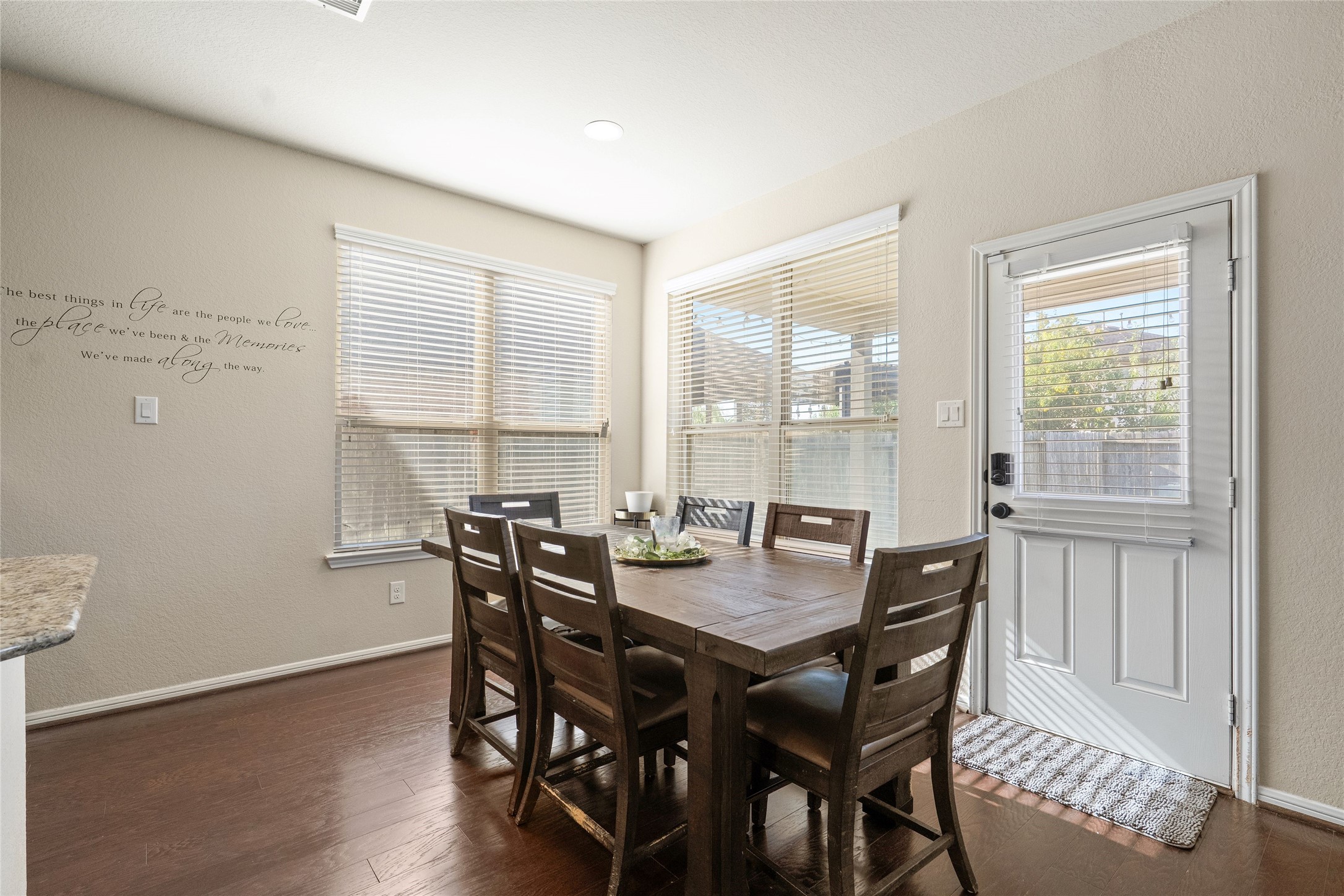 Bright and welcoming dining area with ample natural light, featuring a spacious wooden table and easy access to the outdoor space through a door with charming window blinds.