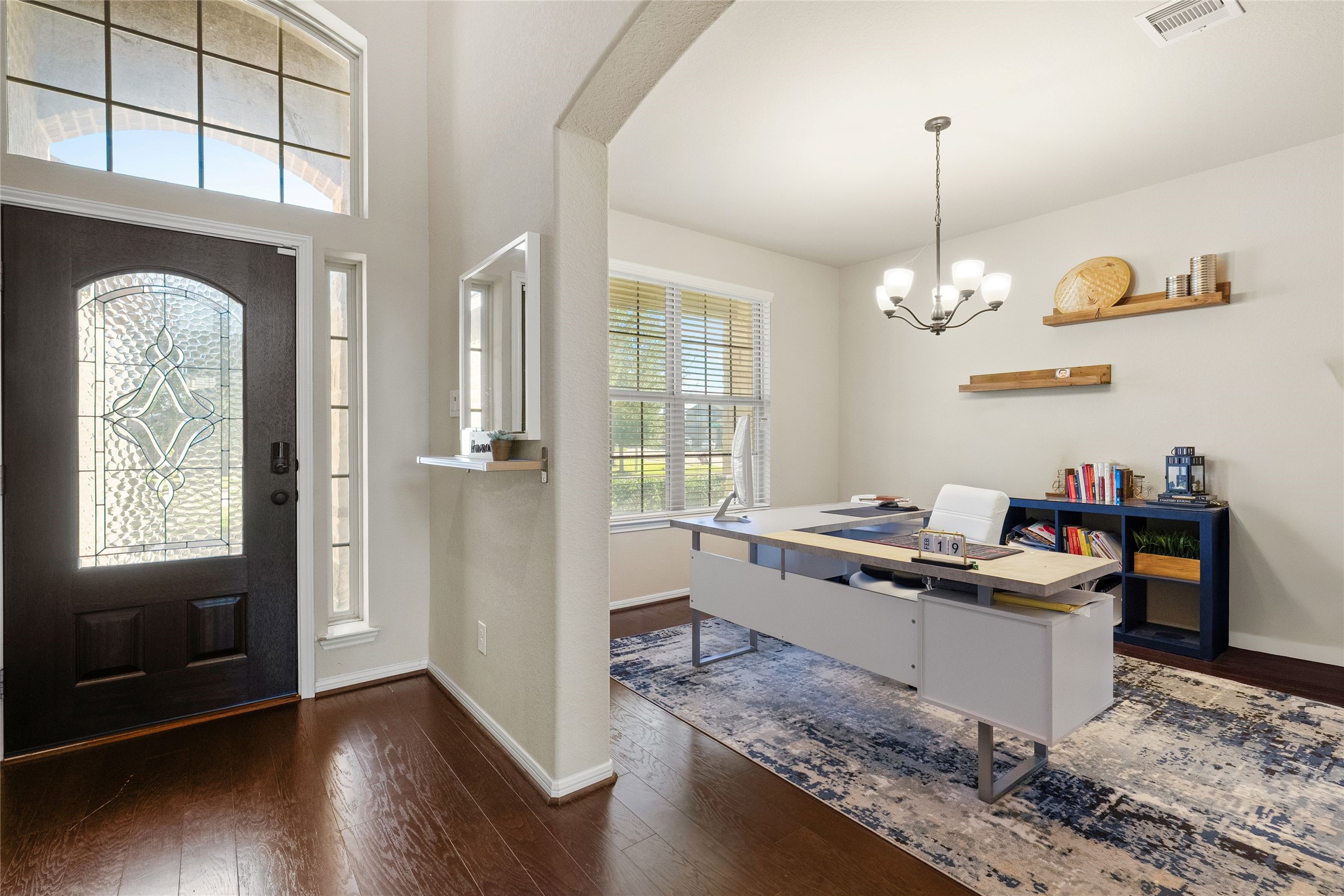 Elegant home office with abundant natural light, featuring a modern desk, stylish chandelier, and built-in shelving, adjacent to a welcoming entryway with a decorative glass door.