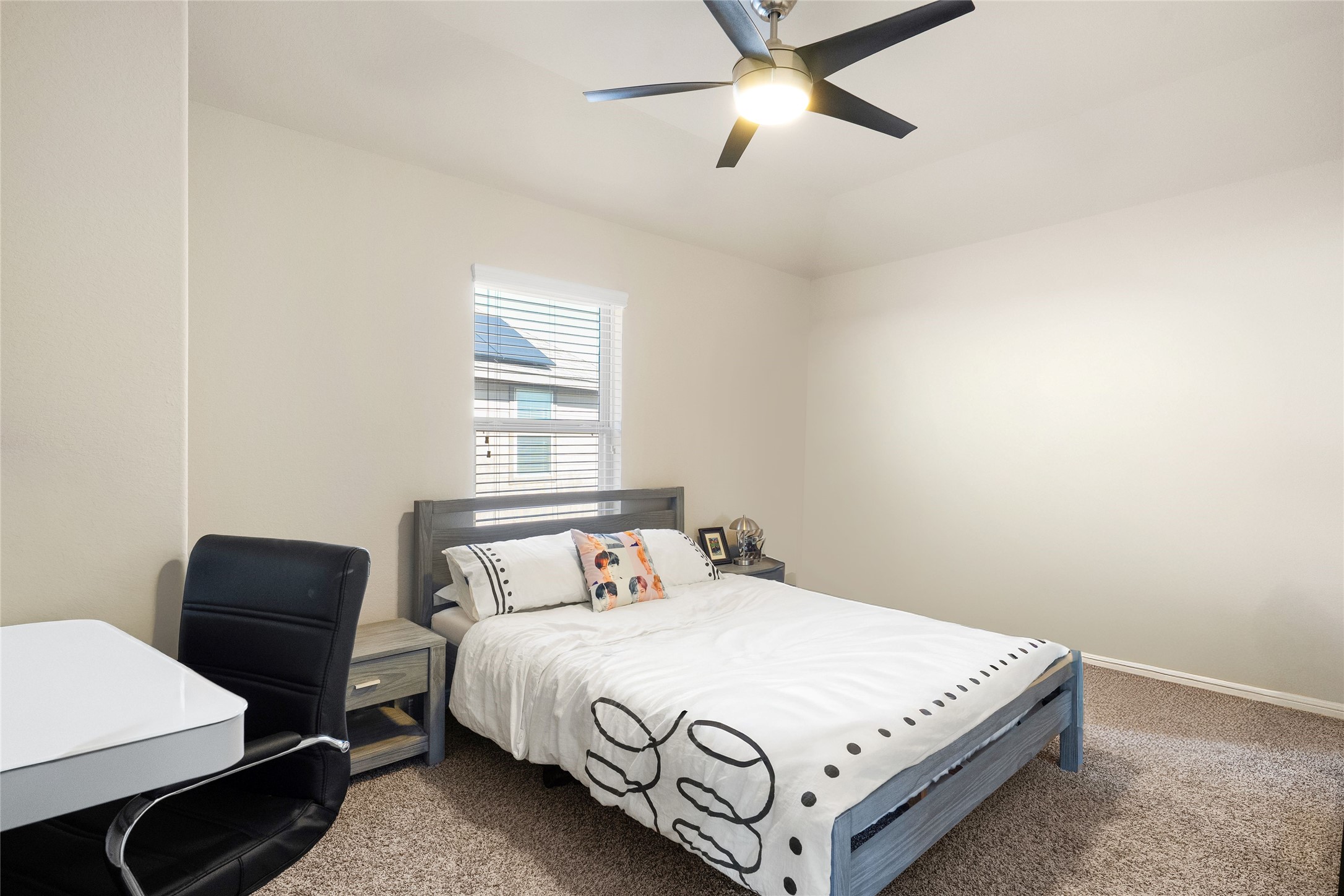 Cozy bedroom with neutral tones features a modern ceiling fan, plush carpeting, and ample natural light through a well-placed window.