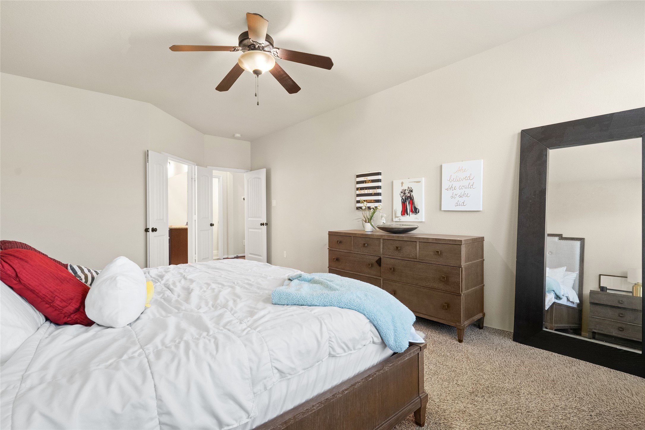Spacious bedroom with plush carpeting, featuring a stylish ceiling fan and ample natural light. The room includes a large mirror, elegant dresser, and inviting double doors leading to an en-suite area.