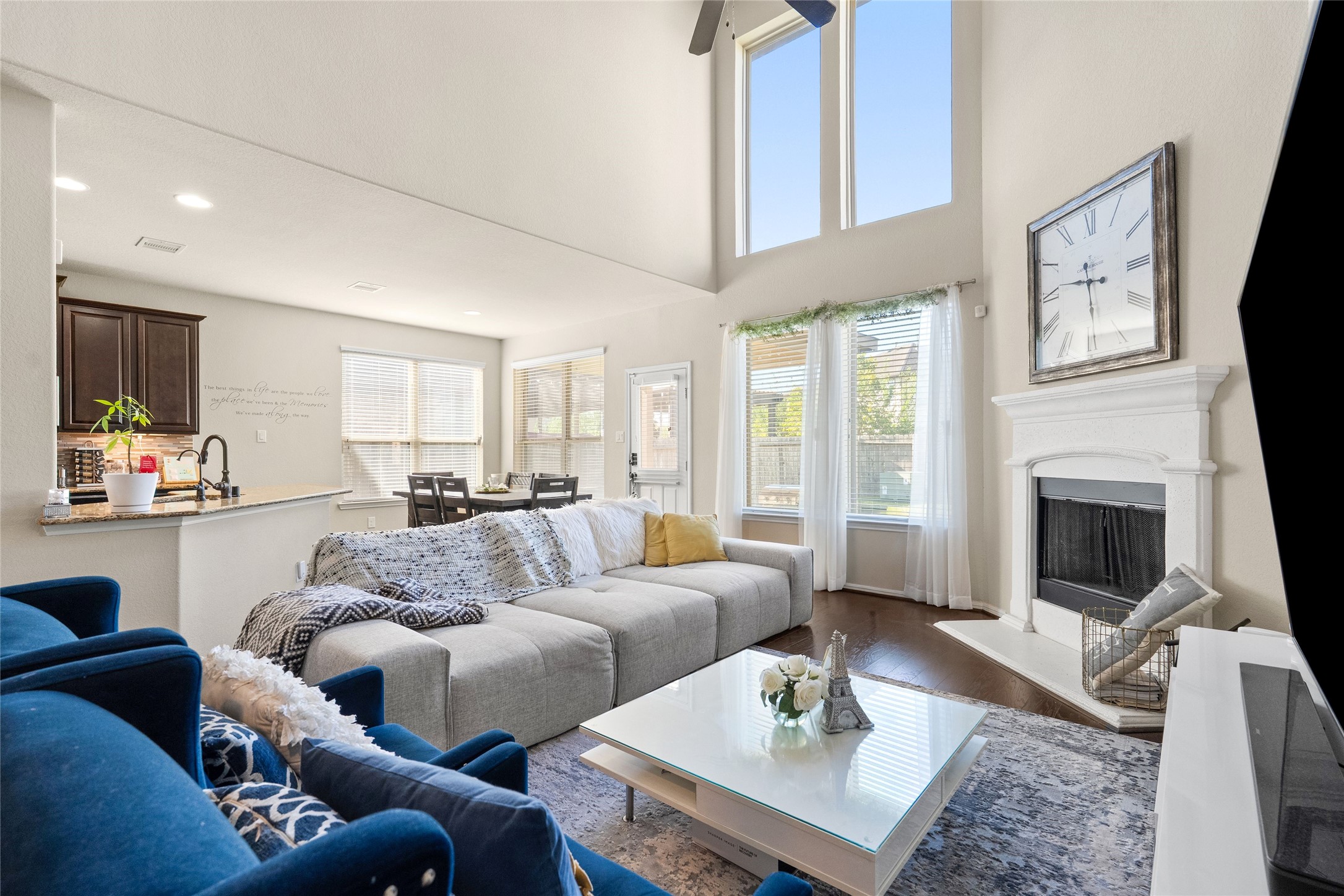 Spacious living room featuring soaring ceilings, abundant natural light from large windows, and a cozy fireplace. The open floor plan flows seamlessly into the dining area and modern kitchen, perfect for entertaining.
