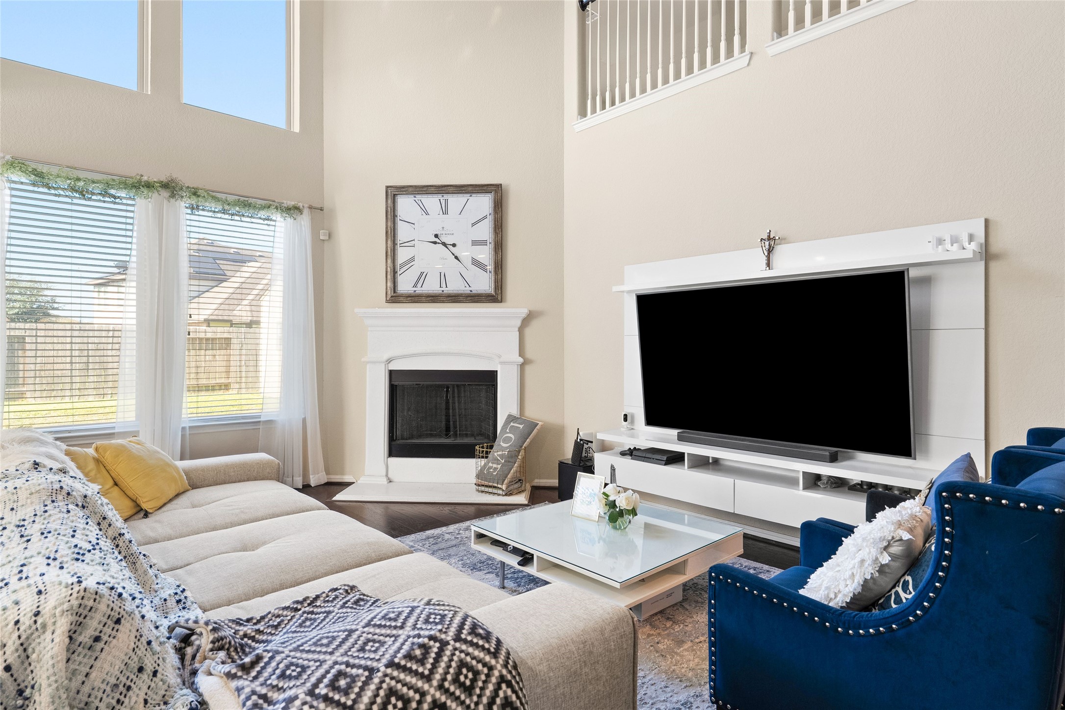 Spacious living room with high ceilings, expansive windows, and plenty of natural light. Features a cozy fireplace, large TV, modern furnishings, and an elegant balcony overlook.