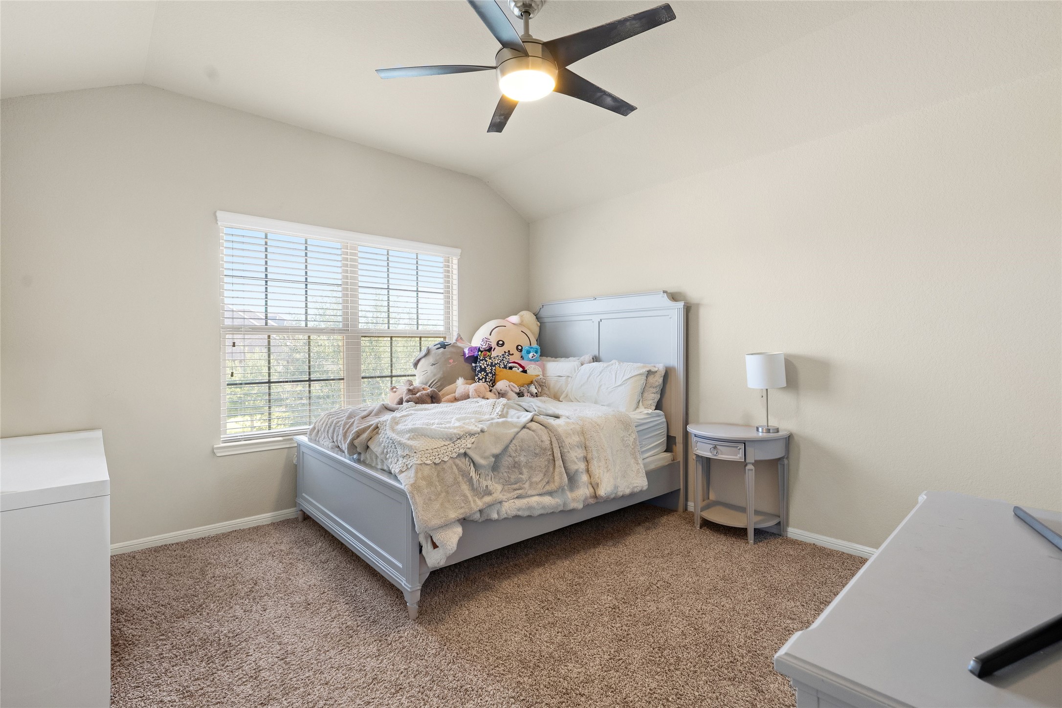 Cozy bedroom with plush carpeting, featuring a large window that bathes the space in natural light, complemented by neutral walls and a modern ceiling fan.