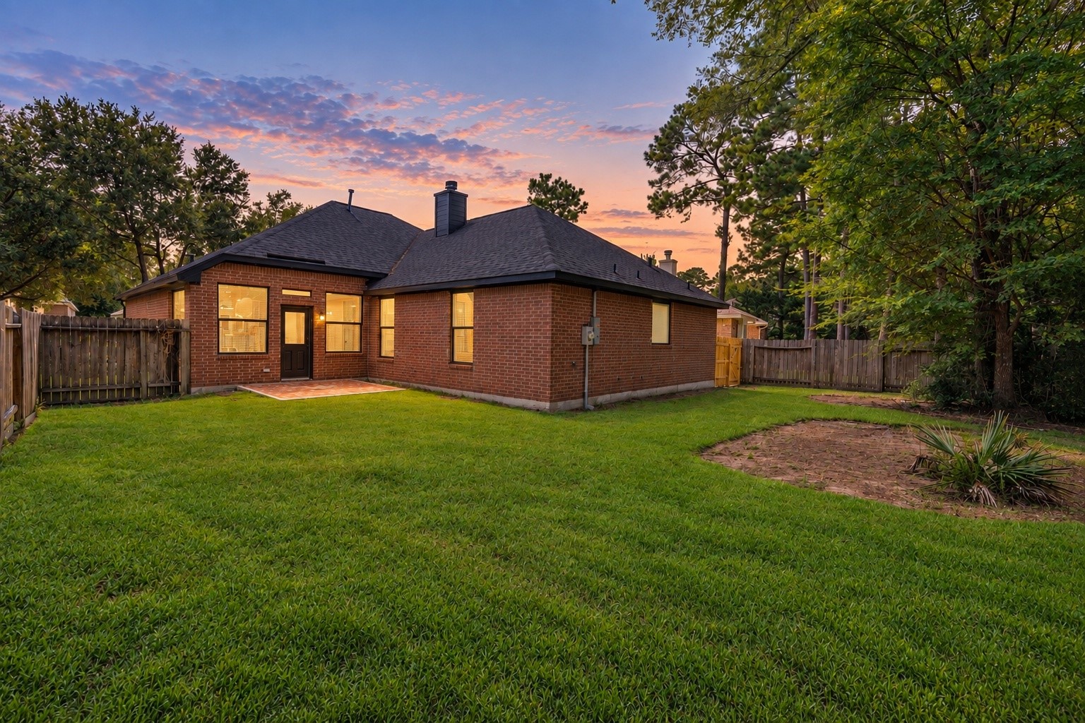 A stunning twilight view of the rear exterior of 18 Griffin Hill Court reveals the home's beautiful brick construction bathed in warm orange and pink sunset hues. Mature trees create a natural privacy canopy around the backyard, while the glow of the evening sky sets a peaceful and magical scene for this remarkable home. (PHOTO MAY BE AI ENHANCED.)