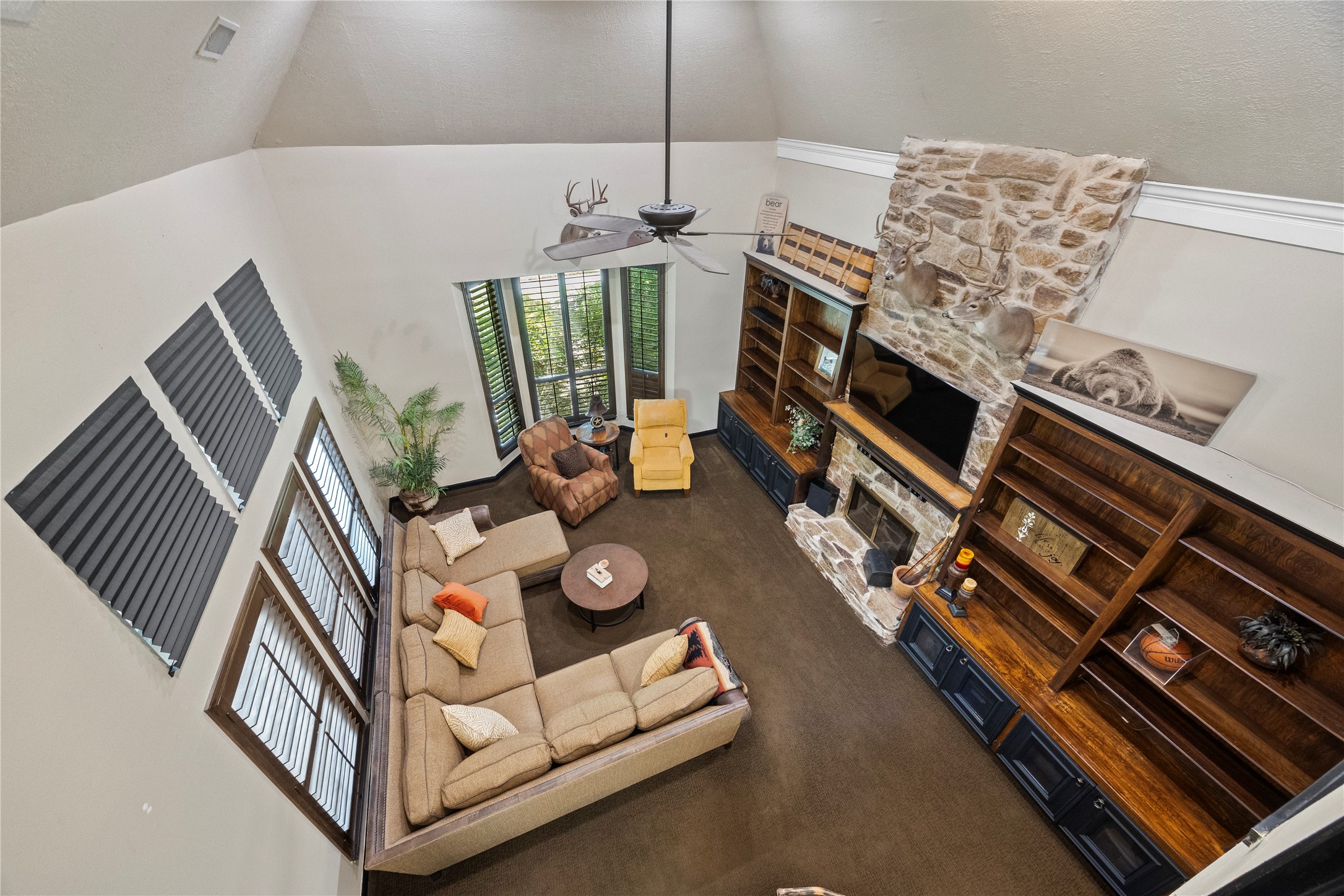 Overhead view of the two-story living room showcases the dramatic ceiling height, floor-to-ceiling stone fireplace, custom built-ins, and expansive windows. The open design creates a connection between the upstairs game room and main living area below.