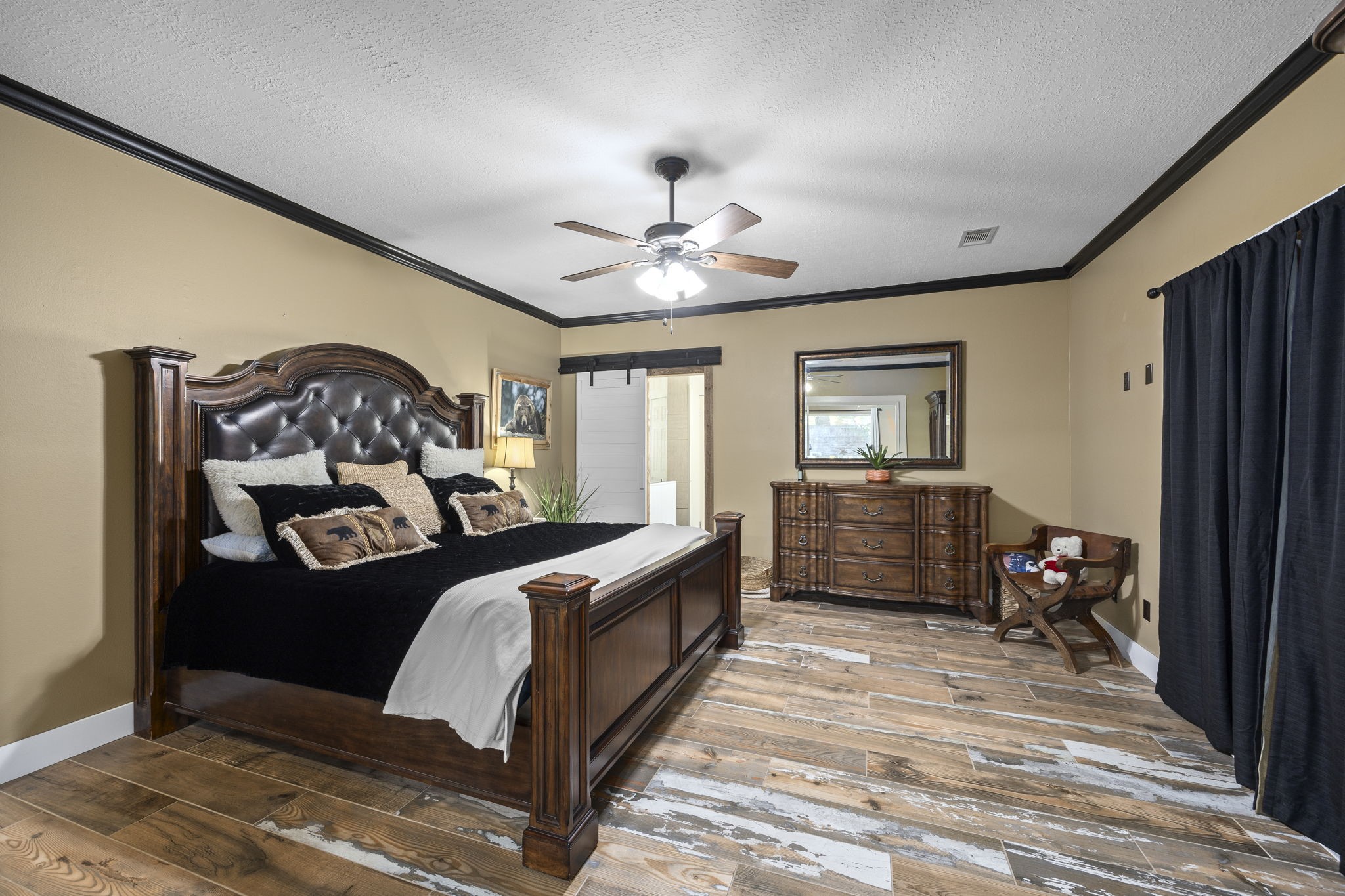 The spacious primary bedroom offers a comfortable retreat with a tray ceiling, wood-look flooring, and plenty of room for substantial furnishings. A neutral color palette and contrasting dark trim add definition to the space.