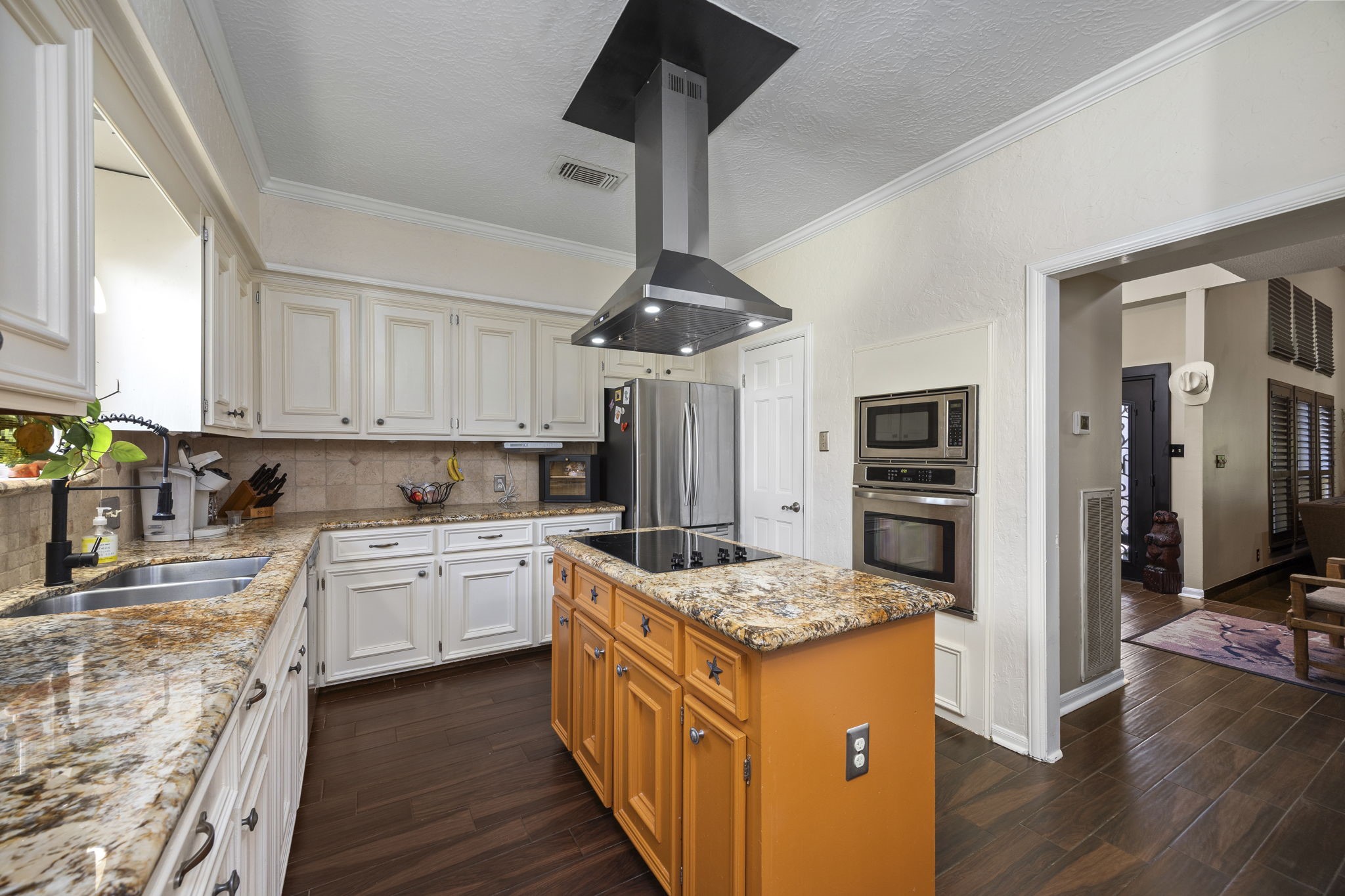 The kitchen features white cabinetry, granite countertops, a contrasting wood island with cooktop and contemporary vent hood, stainless steel appliances, and built-in double ovens. An open doorway provides convenient access to the main living area.