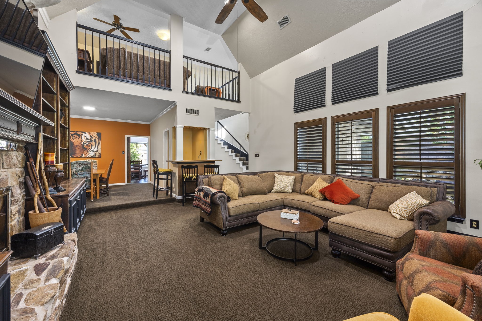 This perspective showcases the living room's impressive scale, with a wall of windows fitted with plantation shutters, an open balcony above, and convenient access to the adjoining dining area and staircase.