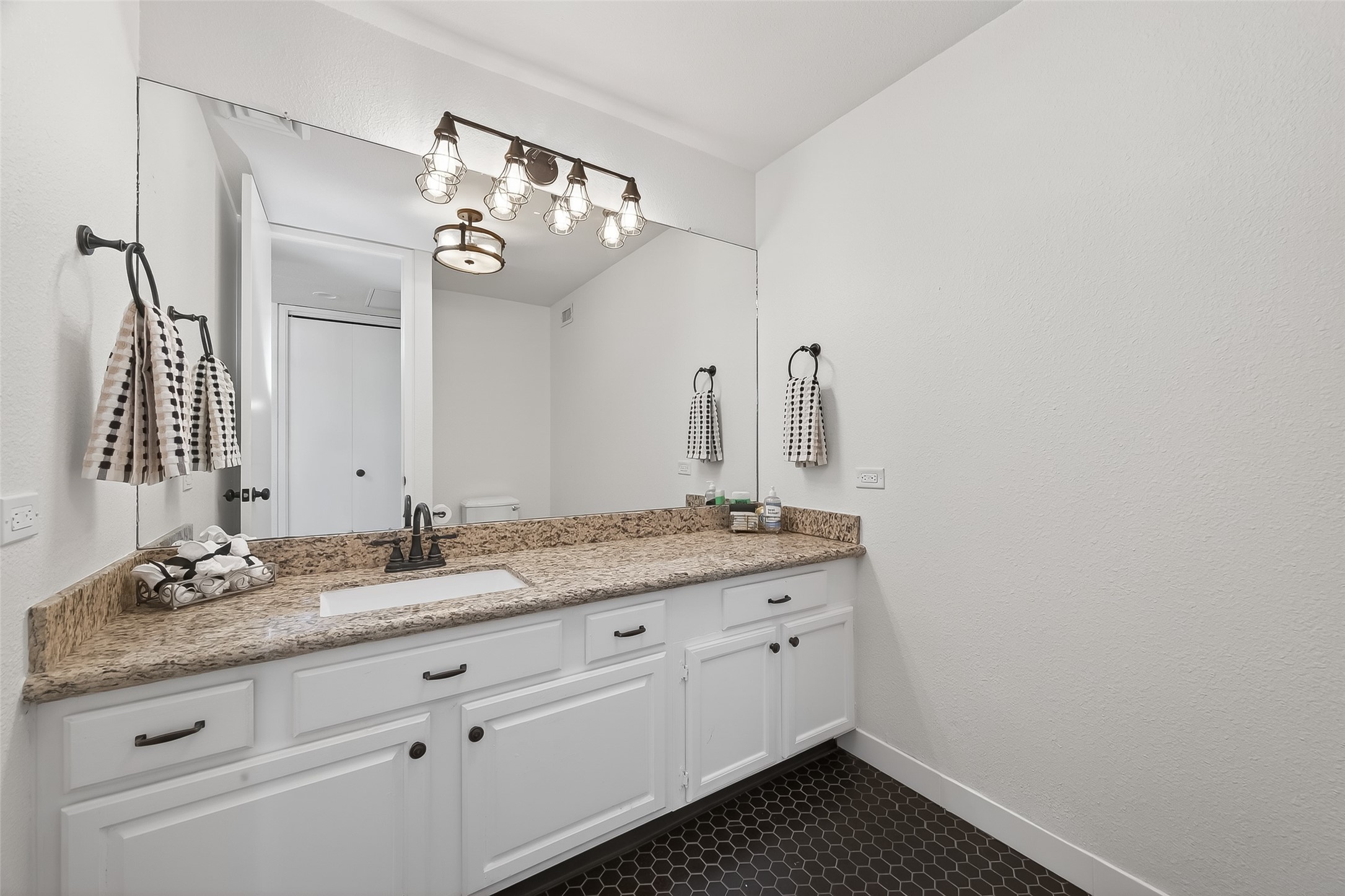 Elegant bathroom featuring a spacious granite countertop with ample cabinet storage, accented by chic lighting and a large mirror, creating a bright and inviting space.