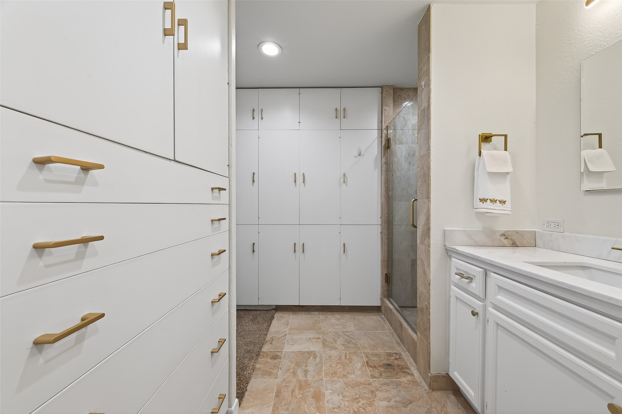 Elegant bathroom featuring ample storage with sleek white cabinetry and brass hardware, complemented by a luxurious marble countertop and a glass-enclosed shower.