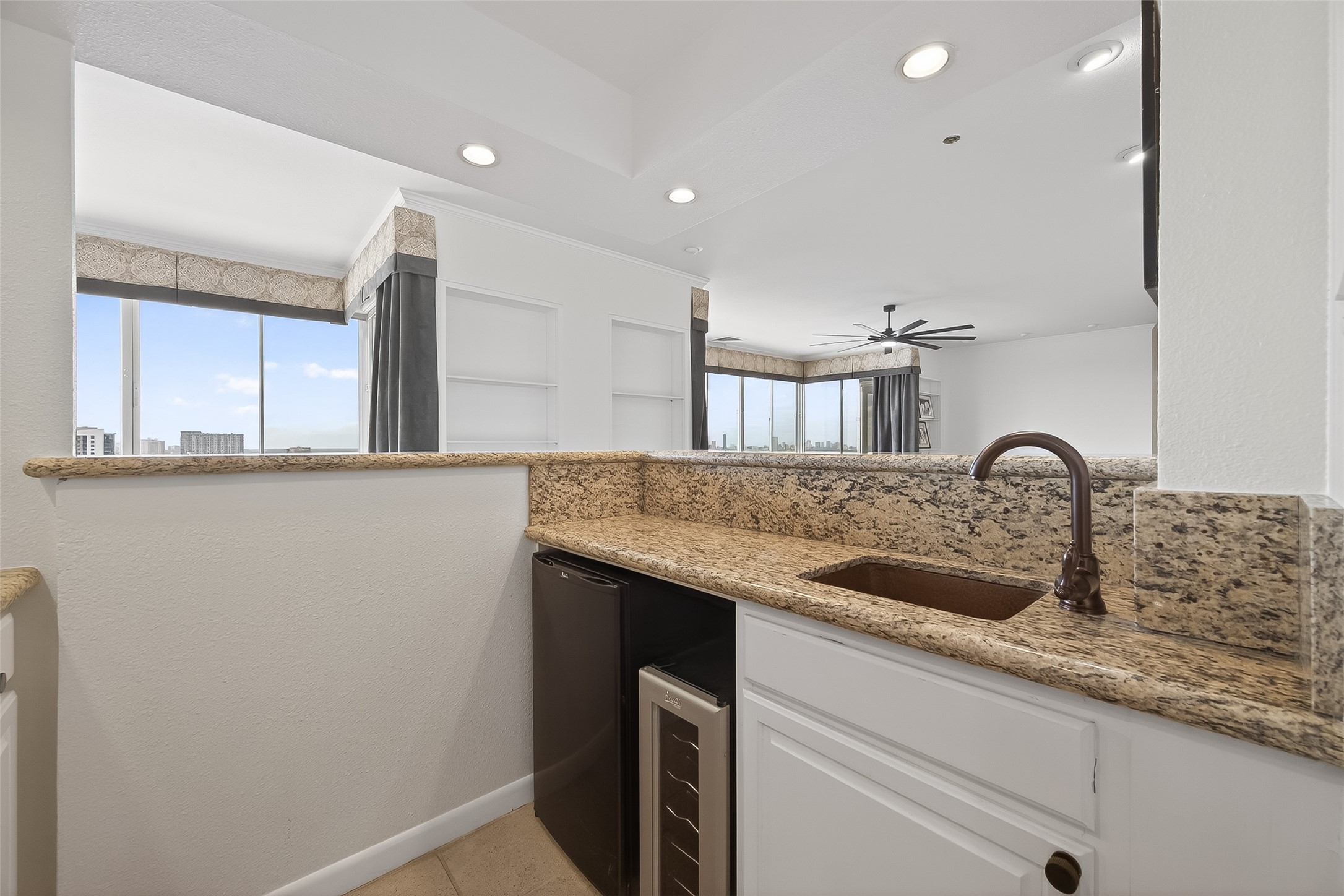 Stylish wet bar area featuring elegant granite countertops, a sleek under-counter wine fridge, and a modern sink, seamlessly opening to a bright living space with expansive city views.