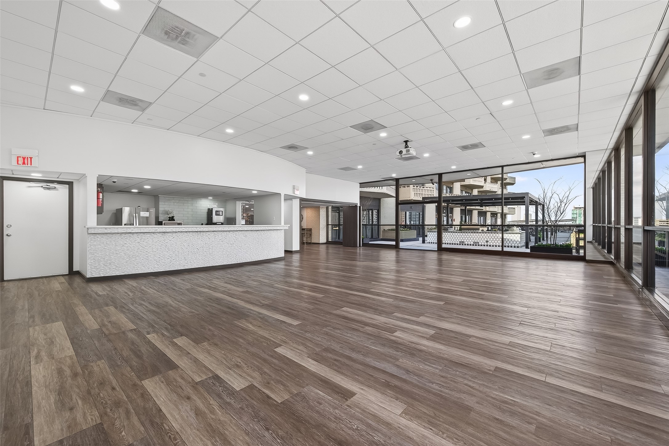 Spacious multi-purpose room with sleek hardwood floors, modern ceiling lighting, and expansive glass windows offering abundant natural light. Features an open kitchen area with a contemporary design, perfect for entertaining or events.
