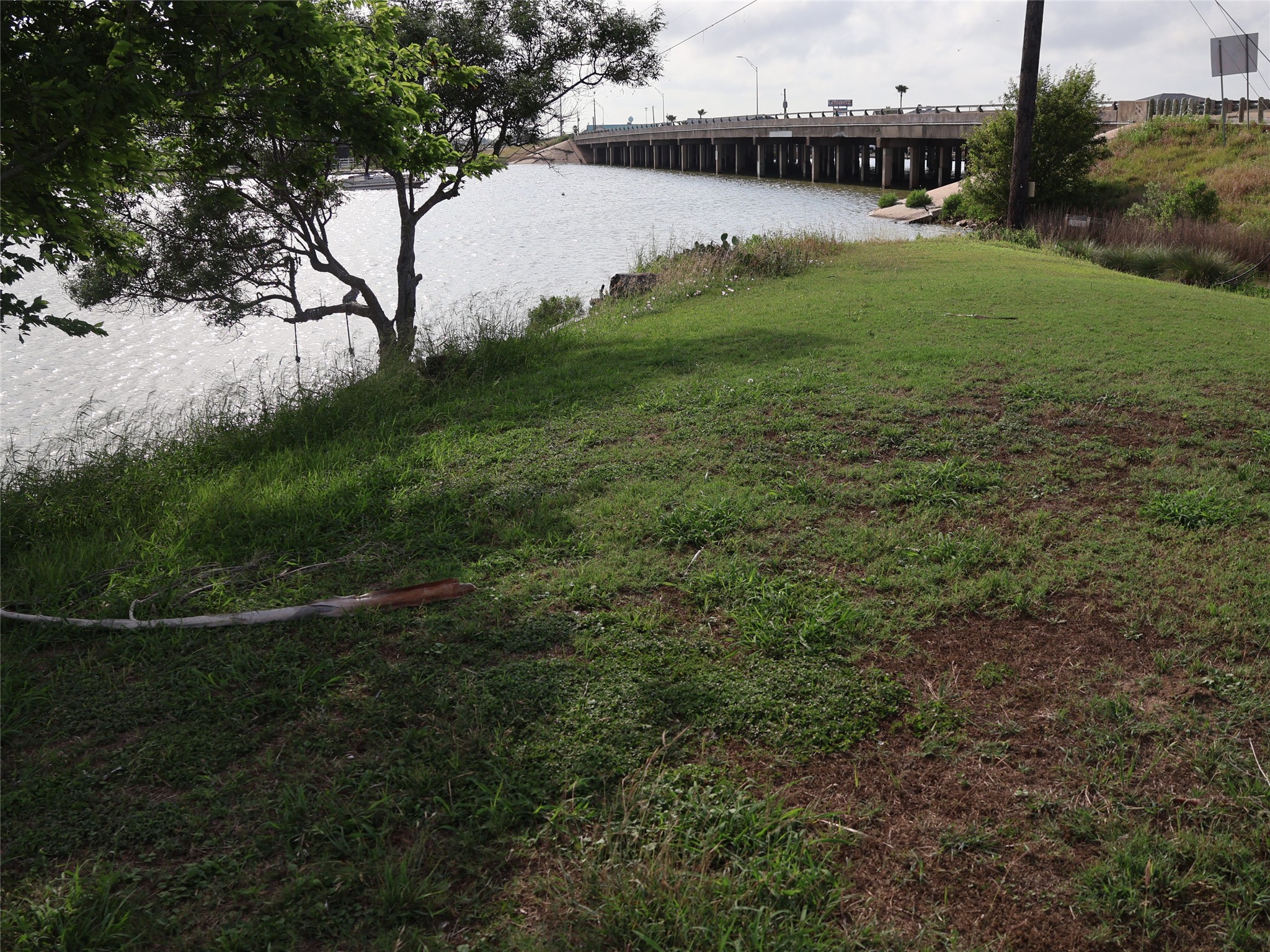 This photo shows a grassy area by a body of water, with a tree providing some shade. In the background, there's a bridge over the water, suggesting easy access to nearby areas. Ideal for those who enjoy waterfront views and outdoor space.