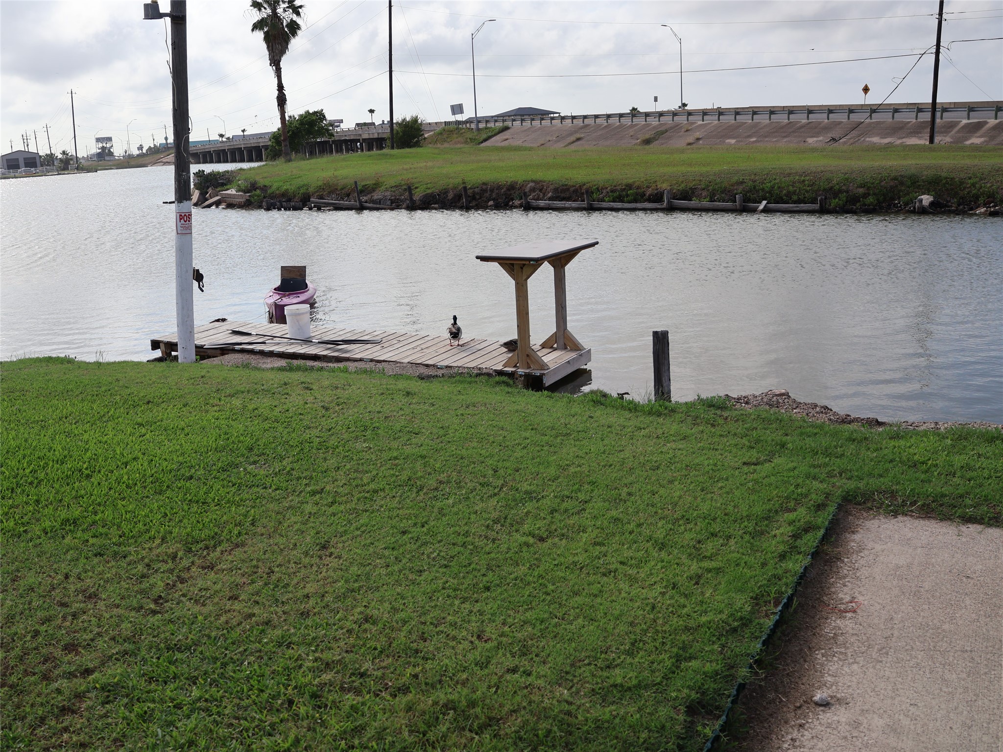 This photo shows a serene waterfront view with a small wooden dock on a grassy bank. The area offers a peaceful setting with open water and nearby road access. Ideal for enjoying outdoor activities and relaxation by the water. This photo shows a serene waterfront view with a small wooden dock on a grassy bank. The area offers a peaceful setting with open water and nearby road access. Ideal for enjoying outdoor activities and relaxation by the water.