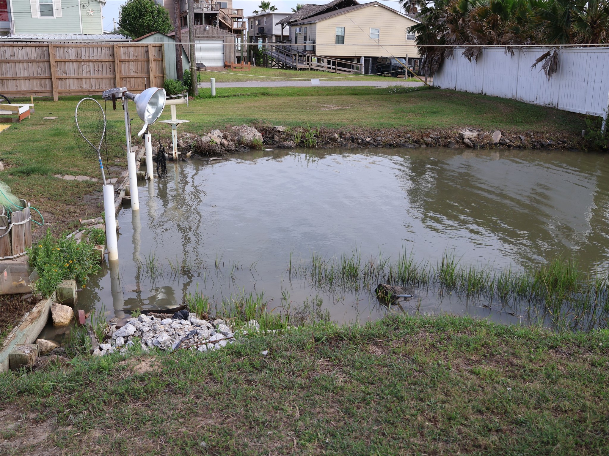 This photo shows a backyard with a small waterway, surrounded by grass and a wooden fence. There's a light fixture and a net nearby, allowing for fishing activities. This photo shows a backyard with a small waterway, surrounded by grass and a wooden fence. There's a light fixture and a net nearby, allowing for fishing activities.