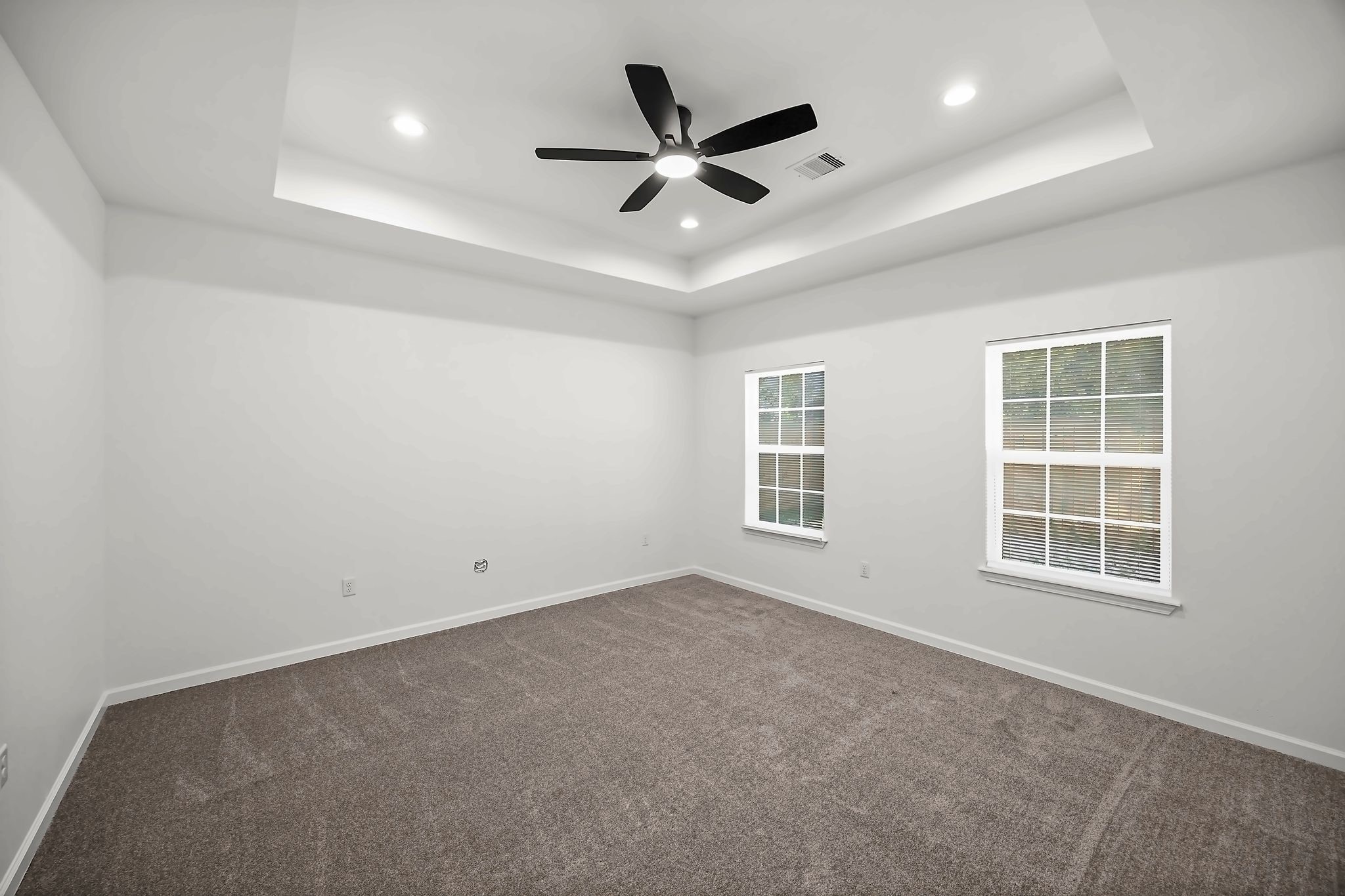 Spacious room featuring a tray ceiling, modern ceiling fan, and plush carpeting, illuminated by recessed lighting and natural light from two large windows.