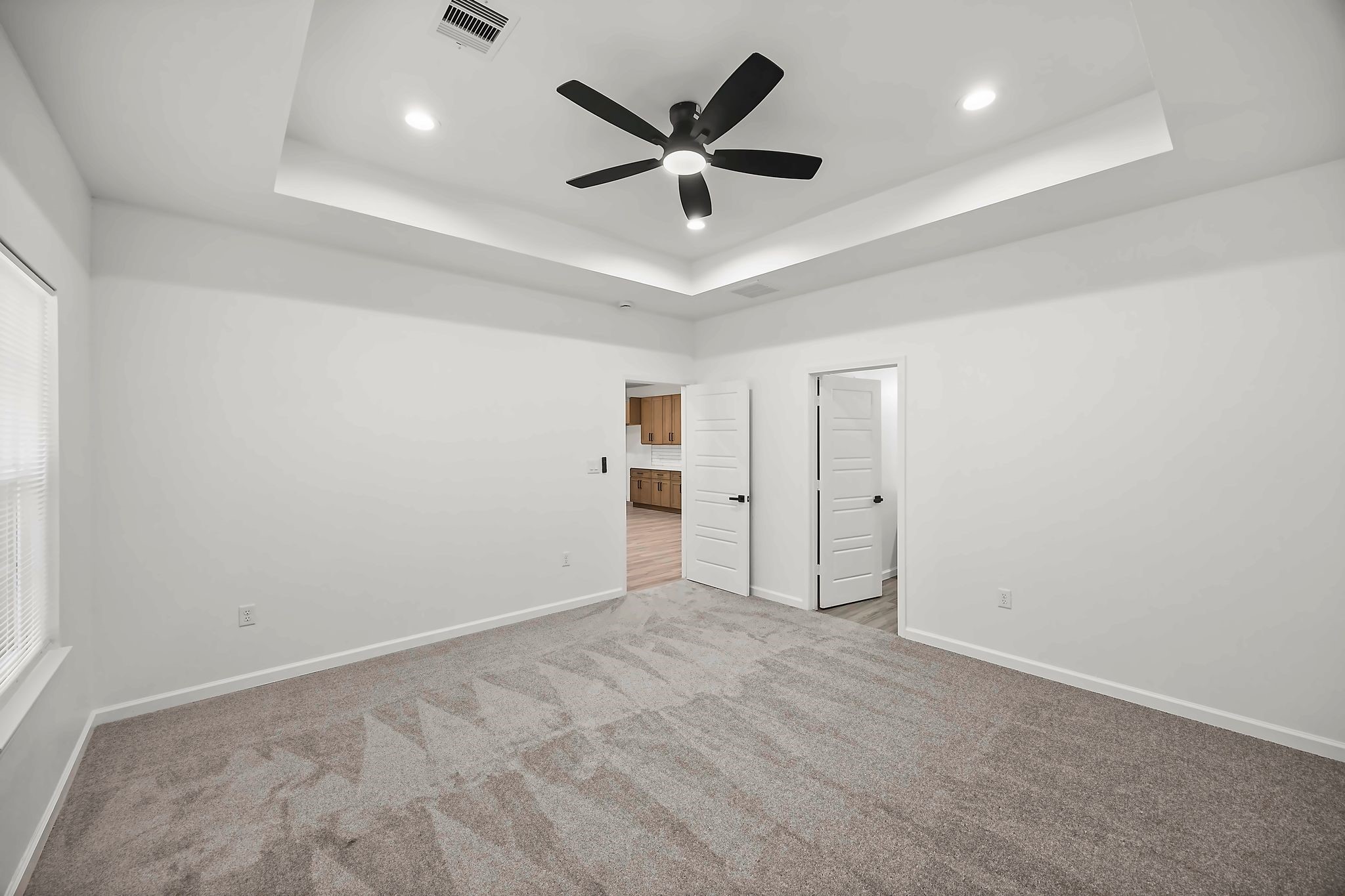 Spacious bedroom with plush carpeting, a modern ceiling fan, and elegant tray ceiling.