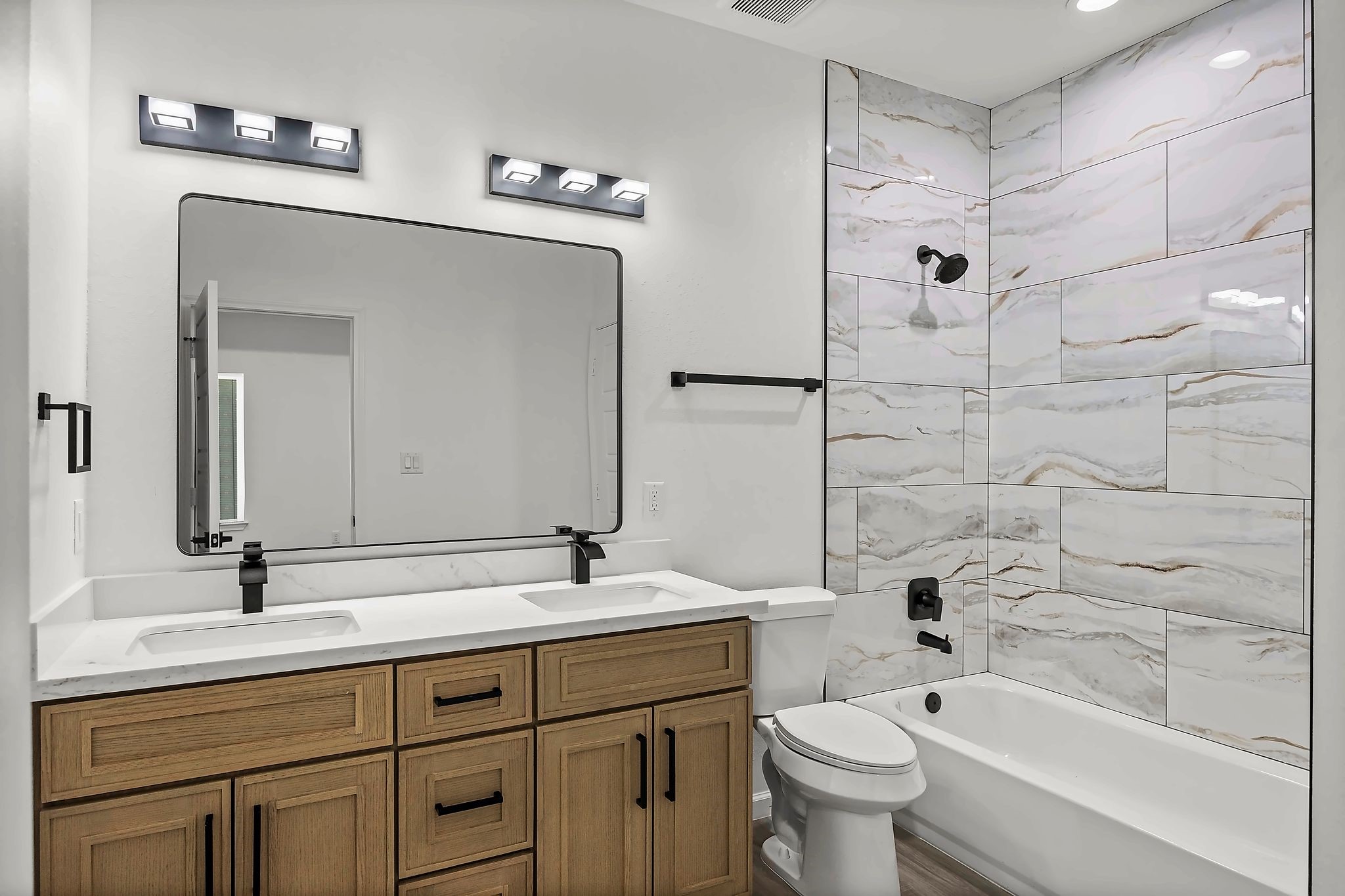 Modern bathroom featuring a sleek double vanity with dark fixtures, elegant marble-tiled shower, and warm wood cabinetry for a touch of luxury.