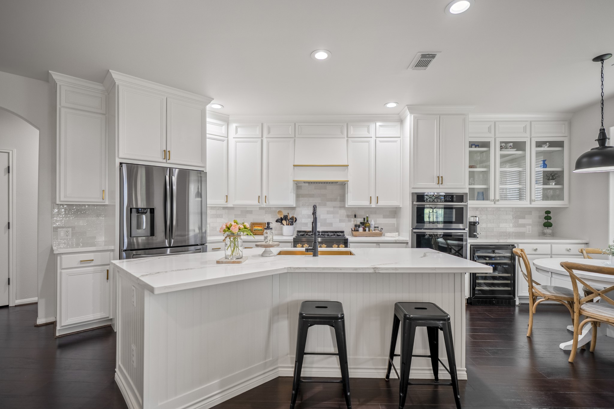 Crisply painted white cabinetry extends to the ceiling, perfectly accented by brushed gold hardware, glass fronted display cabinetry above a beverage station, and modern tile backsplash, with custom vent hood cover.
