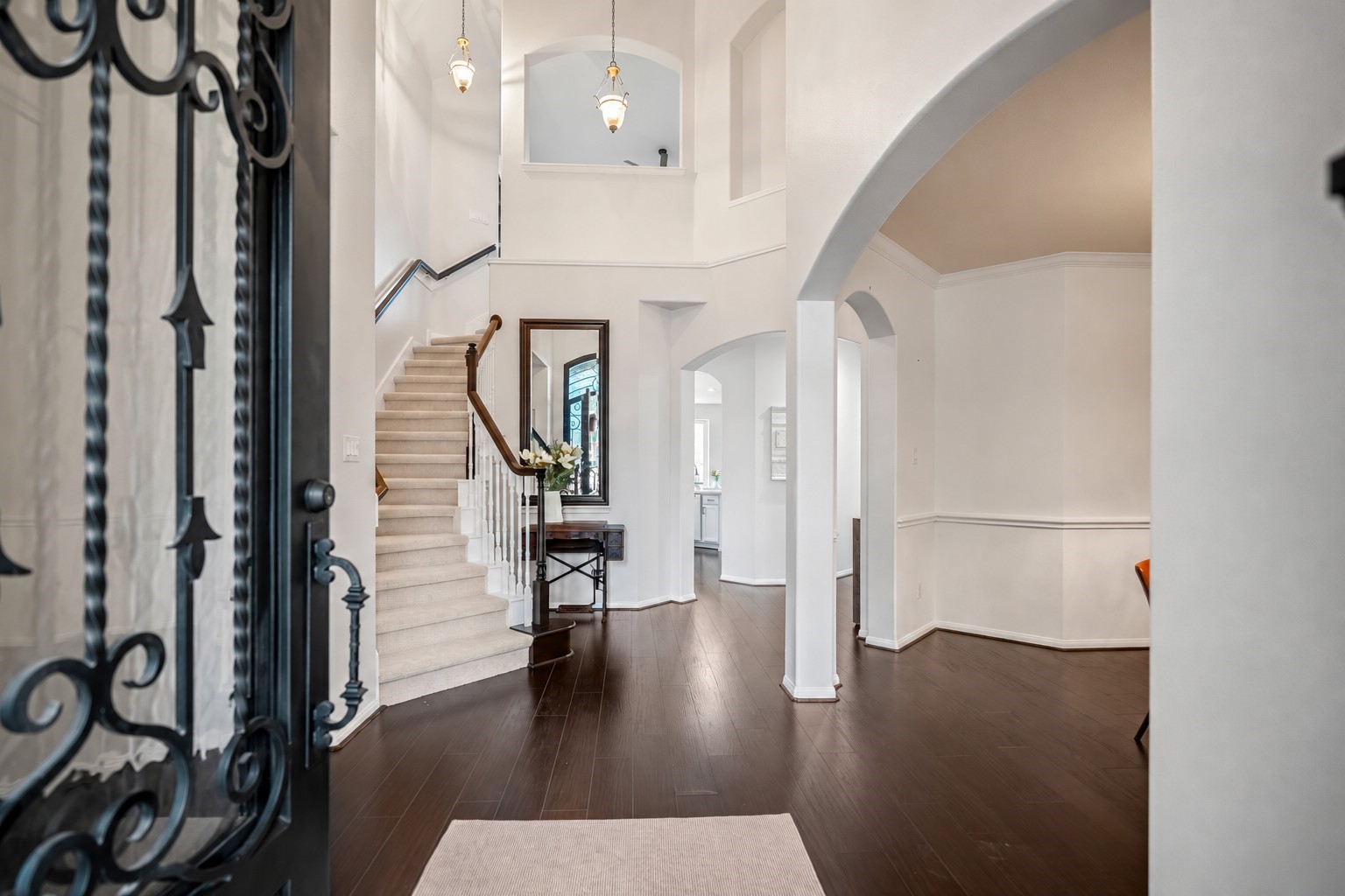 Stunning ornate wrought-iron glass doors open into a towering foyer detailed with elegant architectural arches and gleaming wood look flooring.